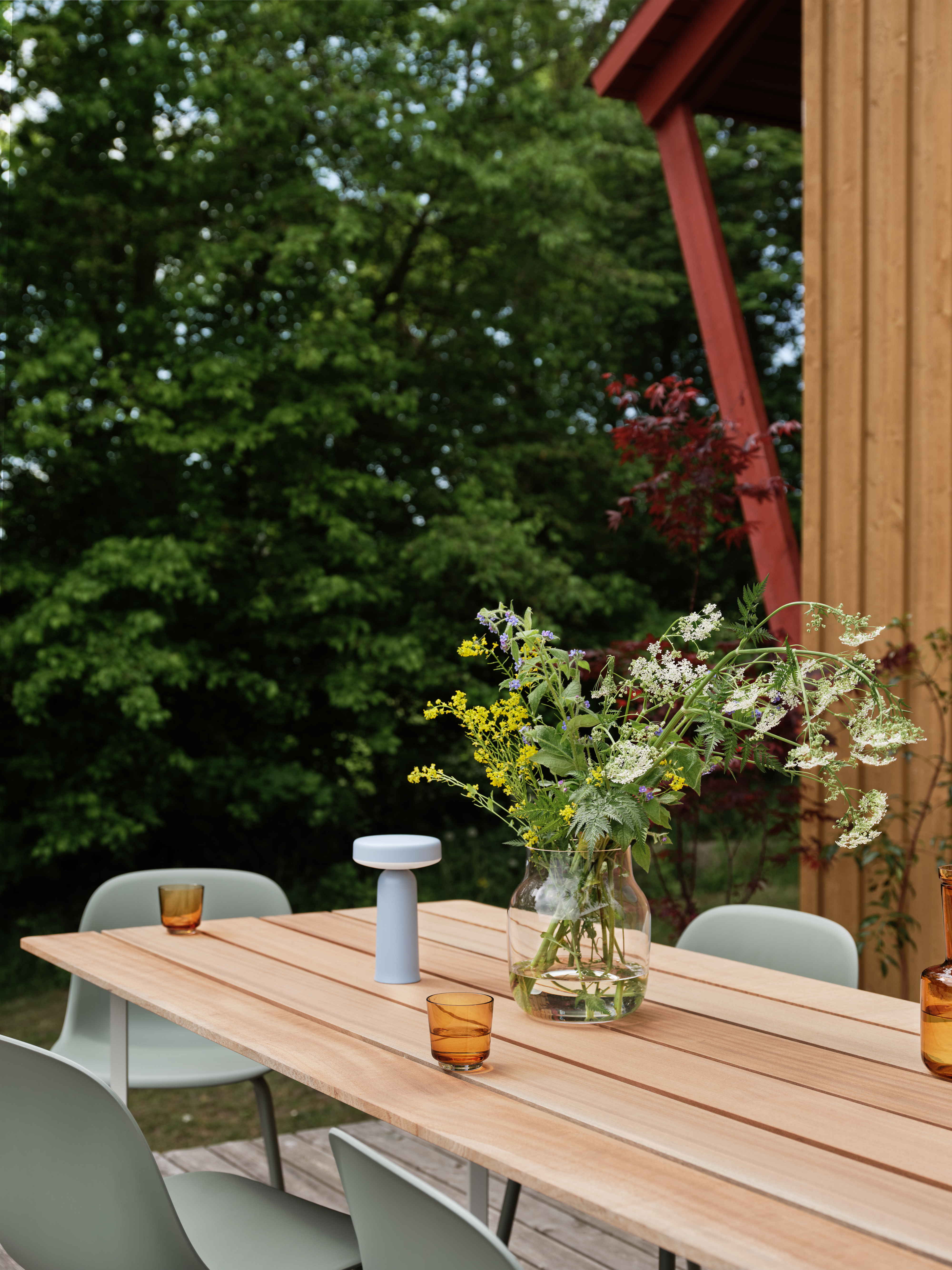 A wooden outdoor dining table with sage green chairs sits on a deck, surrounded by lush greenery. An Ease Lamp and a glass vase with wildflowers decorate the table.