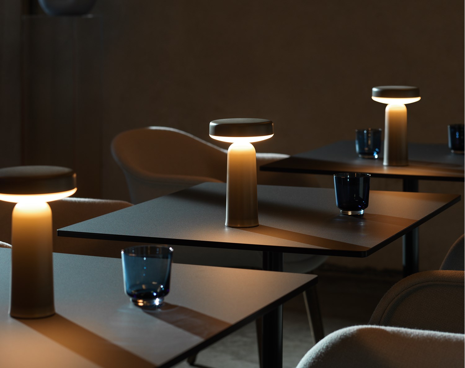A dimly lit café setting with multiple Muuto Ease portable lamps casting a warm glow on dark tables, accompanied by blue drinking glasses and soft chairs.
A dimly lit café setting with multiple Muuto Ease portable lamps casting a warm glow on dark tables, accompanied by blue drinking glasses and soft chairs.