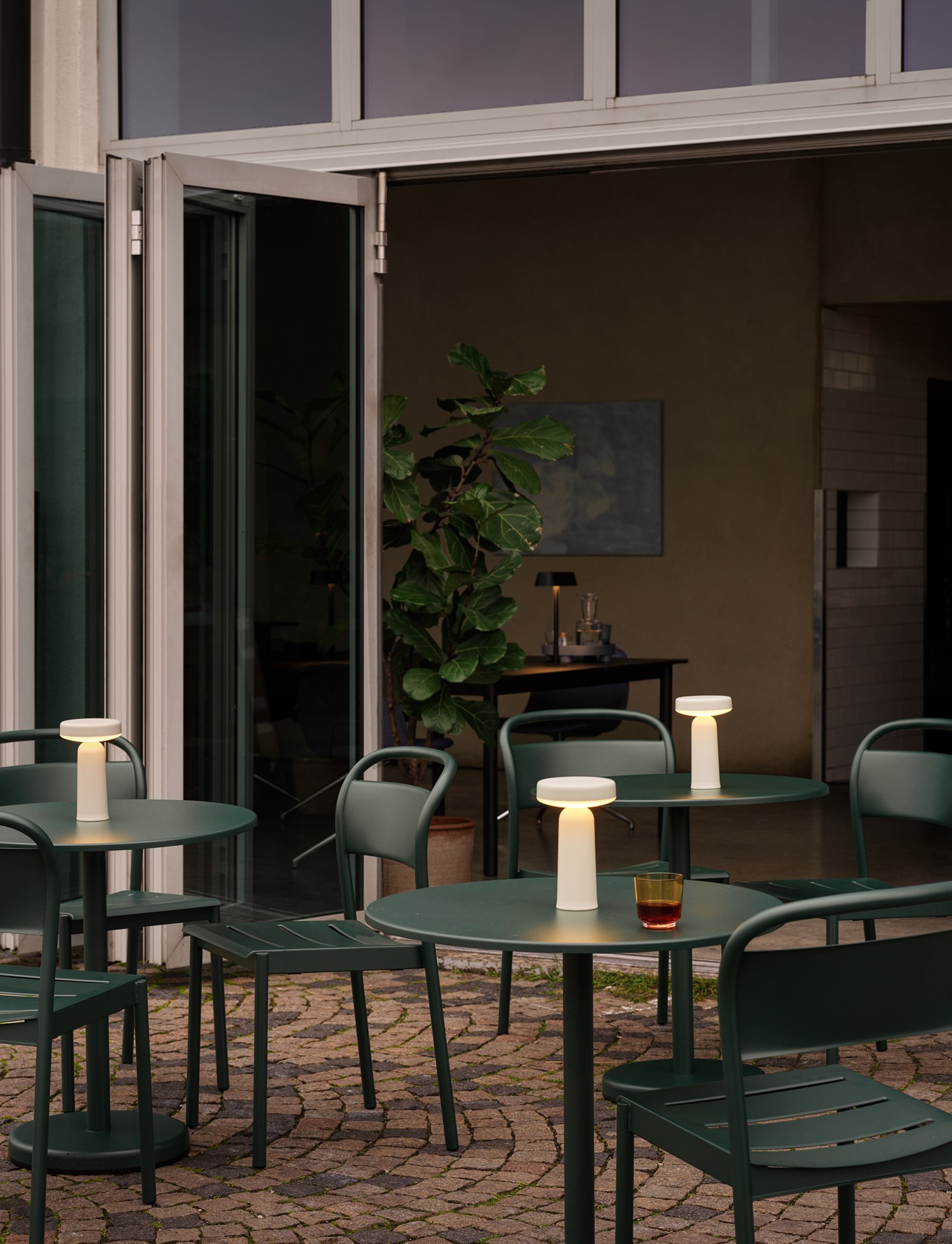 An outdoor café with dark green tables and chairs, each featuring a glowing Ease Lamp. The setting blends indoor and outdoor spaces with open glass doors and potted greenery.
An outdoor café with dark green tables and chairs, each featuring a glowing Ease Lamp. The setting blends indoor and outdoor spaces with open glass doors and potted greenery.