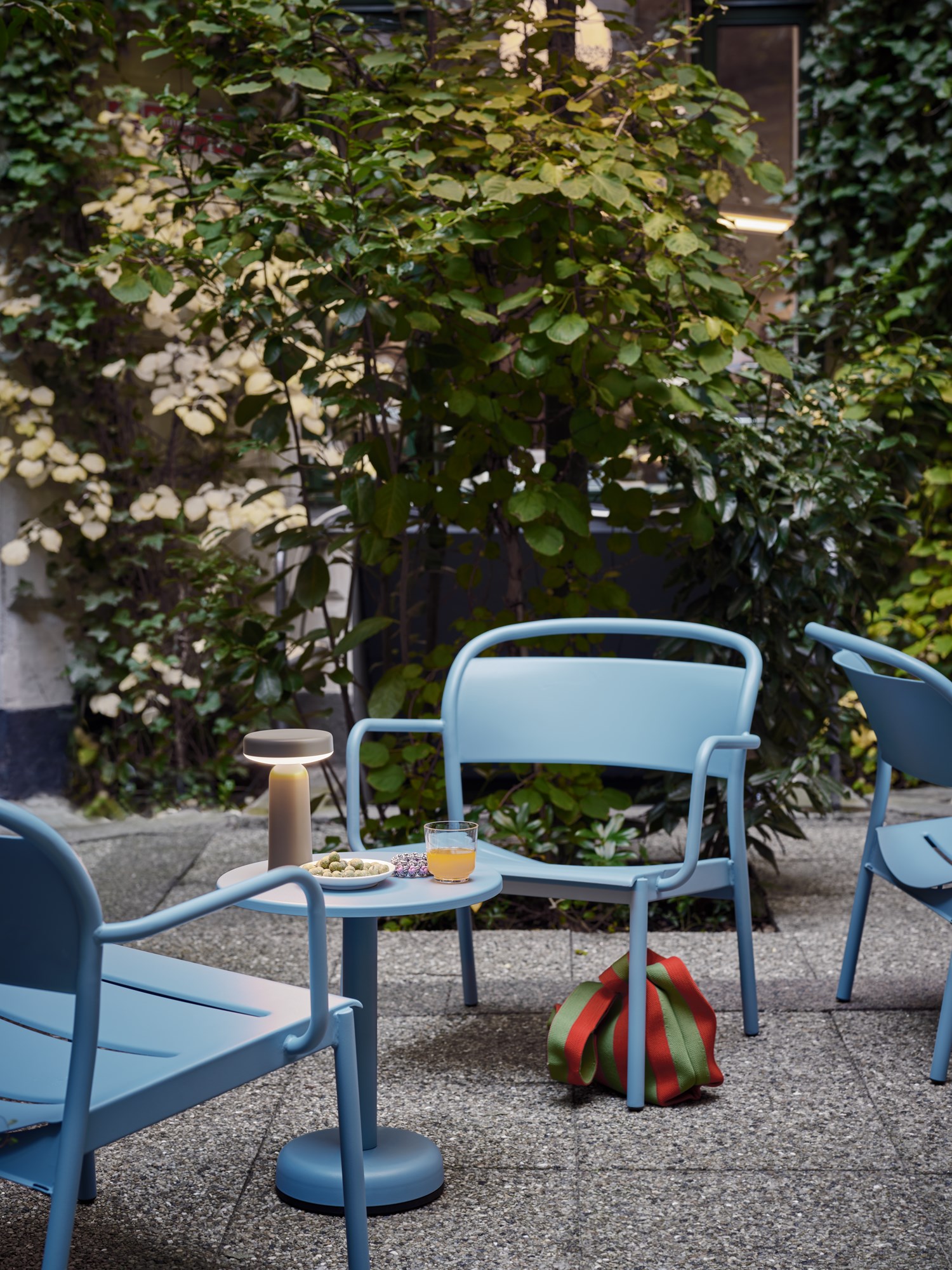 A cozy courtyard setting with light blue metal chairs and a small round table, illuminated by a soft-glowing Ease Lamp. A drink and snacks rest on the table.
A cozy courtyard setting with light blue metal chairs and a small round table, illuminated by a soft-glowing Ease Lamp. A drink and snacks rest on the table.