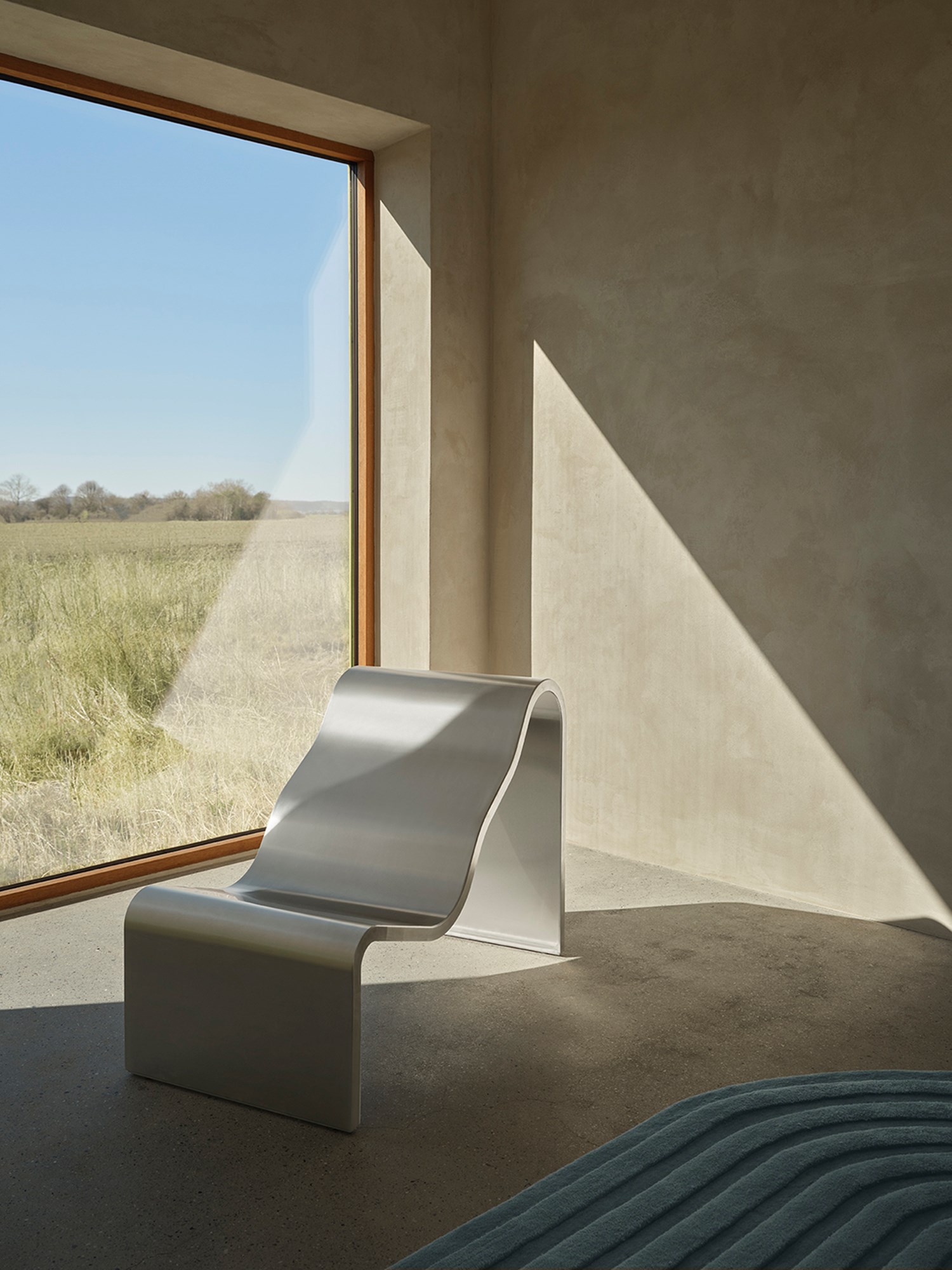 An architectural metal bench with a wavy, sculptural design is placed inside in the sunlight on a Relevo rug in Sage Green. An architectural metal bench with a wavy, sculptural design is placed inside in the sunlight on a Relevo rug in Sage Green.