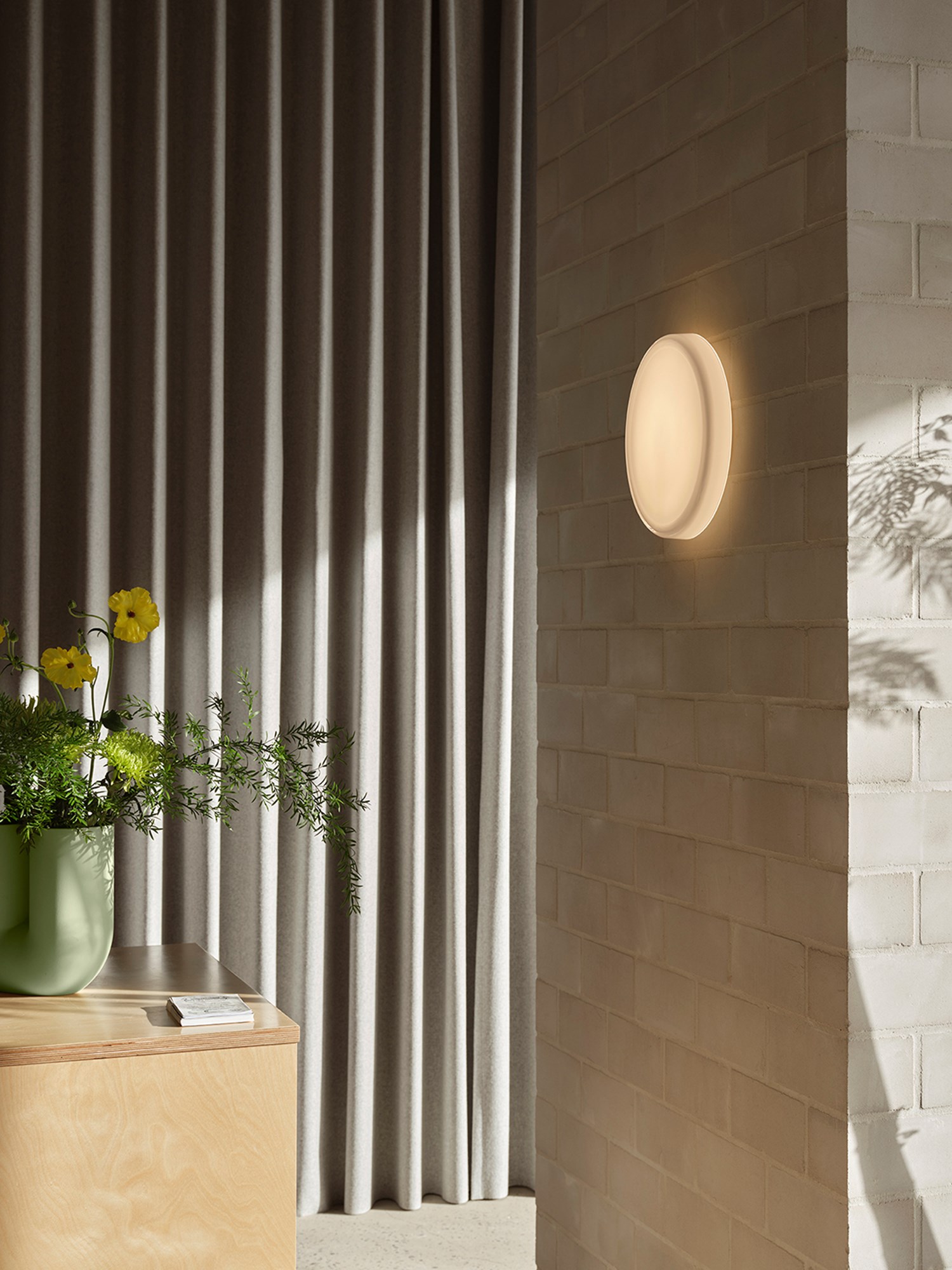 A minimalistich hallway featuring brushed stone walls. A round wall lamp hanging on the wall and a sculptural vase in light green is placed on a wooden table, creating a serene and modern atmosphere. A minimalistich hallway featuring brushed stone walls. A round wall lamp hanging on the wall and a sculptural vase in light green is placed on a wooden table, creating a serene and modern atmosphere.