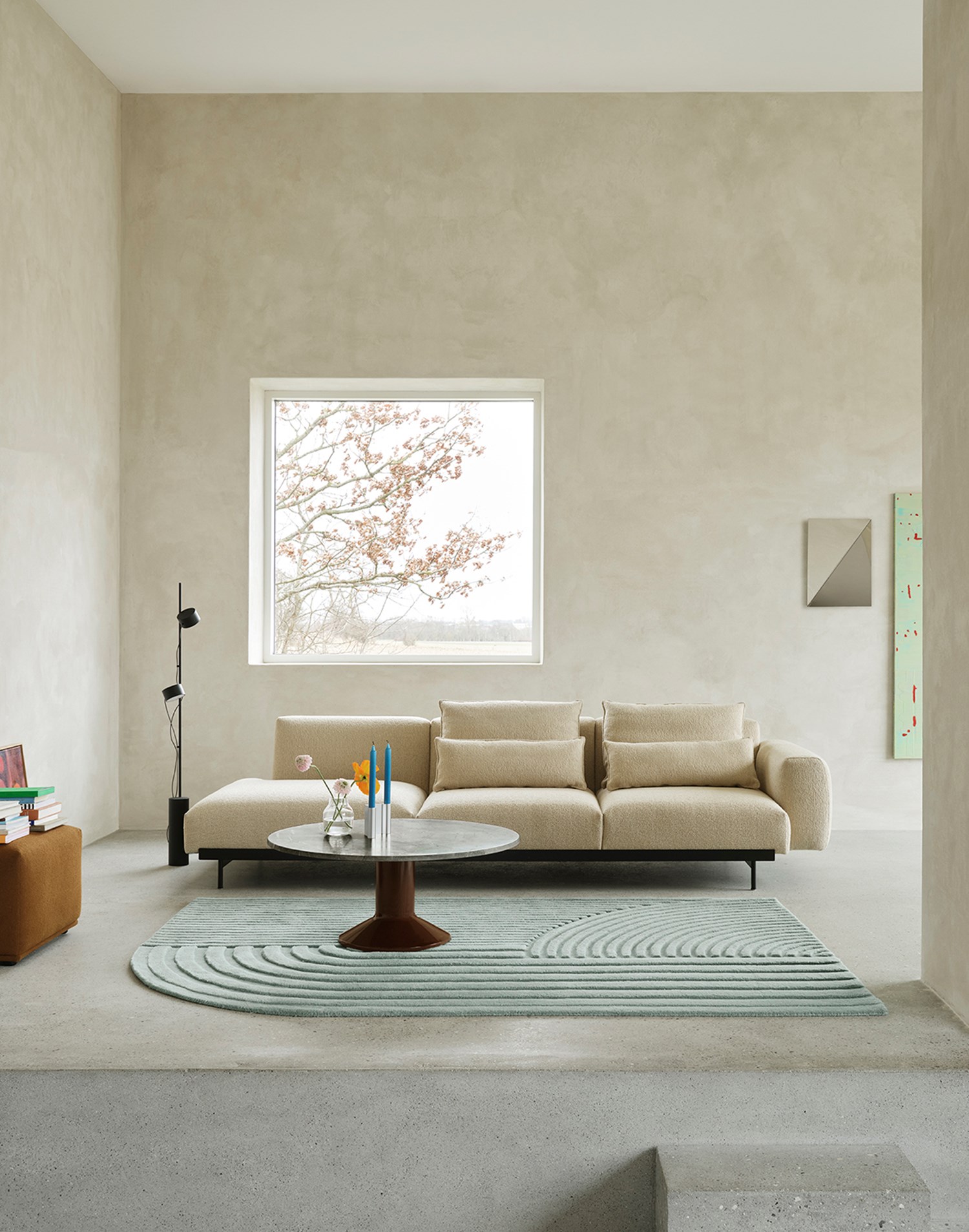A sand-colored sofa is positioned against a brushed grey stone wall. A black floor lamp stands beside the sofa, while a round coffee table in marble and burgundy is placed in front, atop a light blue rug. The arrangement creates a stylish and serene living space. A sand-colored sofa is positioned against a brushed grey stone wall. A black floor lamp stands beside the sofa, while a round coffee table in marble and burgundy is placed in front, atop a light blue rug. The arrangement creates a stylish and serene living space.