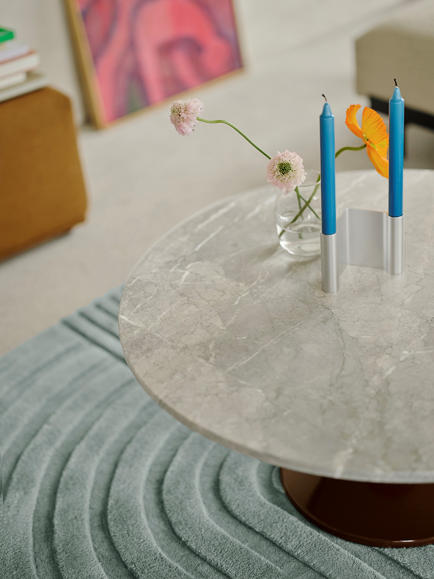 A close-up of a burgundy and marble coffee table, with a modern candle holder and a glass vase with white flowers placed on top. A soft light blue rug is positioned underneath the table, adding warmth and creating a cozy yet contemporary setting. A close-up of a burgundy and marble coffee table, with a modern candle holder and a glass vase with white flowers placed on top. A soft light blue rug is positioned underneath the table, adding warmth and creating a cozy yet contemporary setting.