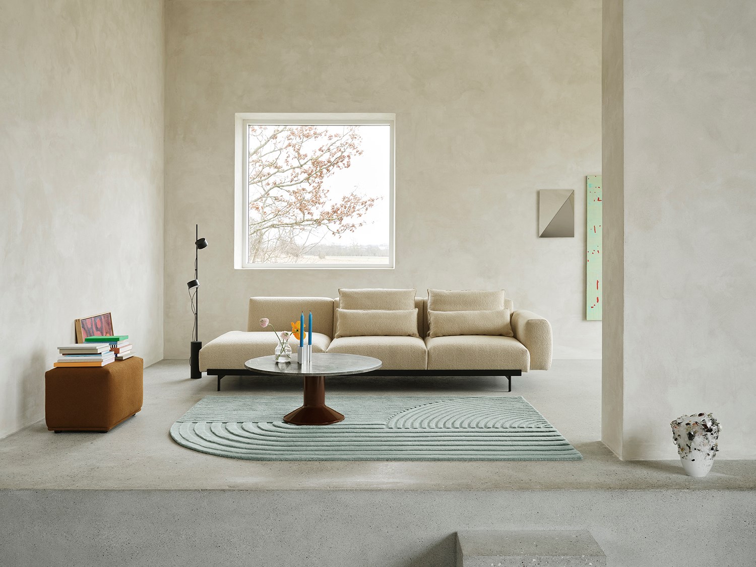 A sand-colored sofa is positioned against a brushed grey stone wall. A black floor lamp stands beside the sofa, while a round coffee table in marble and burgundy is placed in front, atop a light blue rug. The arrangement creates a stylish and serene living space. A sand-colored sofa is positioned against a brushed grey stone wall. A black floor lamp stands beside the sofa, while a round coffee table in marble and burgundy is placed in front, atop a light blue rug. The arrangement creates a stylish and serene living space.