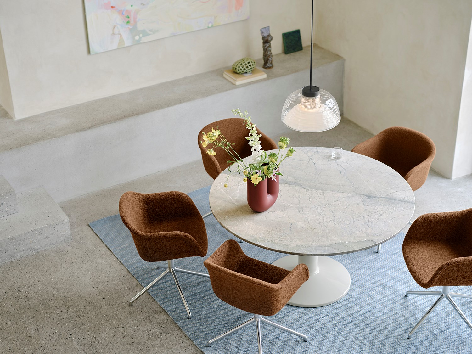 Seen from above, a round grey marble dining table sits in a minimalistic dining room with brushed stone walls and floors. Soft brown dining chairs surround the table, with a light blue rug beneath adding a pop of color. A glass pendant lamp hangs overhead, enhancing the serene and modern atmosphere. Seen from above, a round grey marble dining table sits in a minimalistic dining room with brushed stone walls and floors. Soft brown dining chairs surround the table, with a light blue rug beneath adding a pop of color. A glass pendant lamp hangs overhead, enhancing the serene and modern atmosphere.