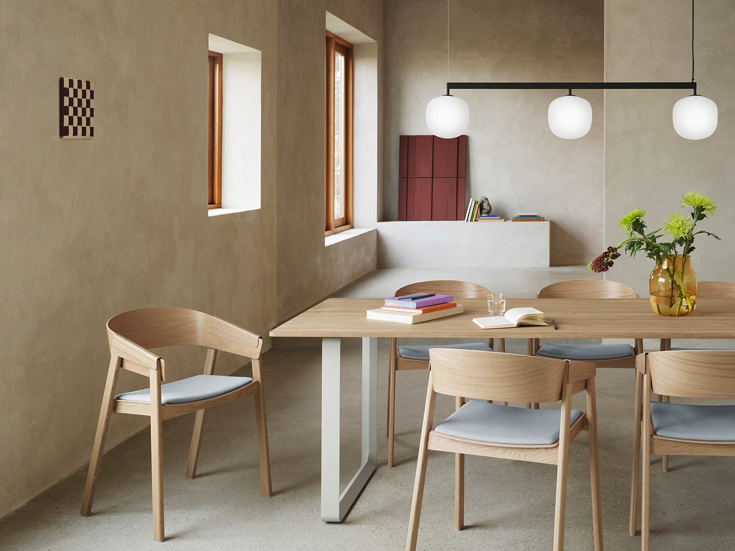 Modern wooden dining chairs are arranged around a wooden dining table in a minimalistic kitchen lounge area with brushed stone surfaces. A sleek, modern pendant lamp hangs above the table setting, enhancing the clean and natural aesthetic. Modern wooden dining chairs are arranged around a wooden dining table in a minimalistic kitchen lounge area with brushed stone surfaces. A sleek, modern pendant lamp hangs above the table setting, enhancing the clean and natural aesthetic.
