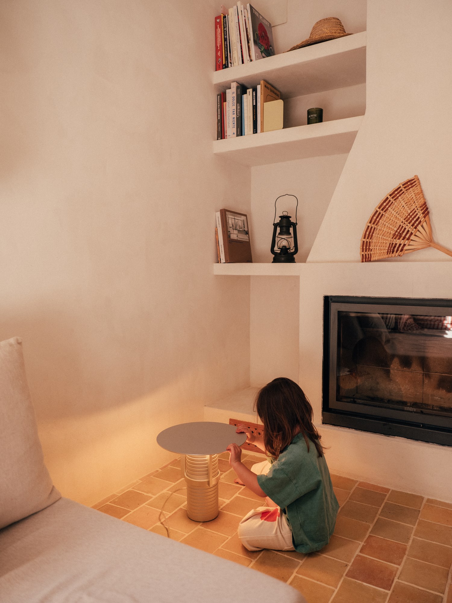 Nuria’s daughter interacting with the Set Lamp. The warm-toned living space features terracotta flooring, a white stucco fireplace, built-in bookshelves, and a beige sofa, evoking a minimalist Mediterranean aesthetic. Nuria’s daughter interacting with the Set Lamp. The warm-toned living space features terracotta flooring, a white stucco fireplace, built-in bookshelves, and a beige sofa, evoking a minimalist Mediterranean aesthetic.