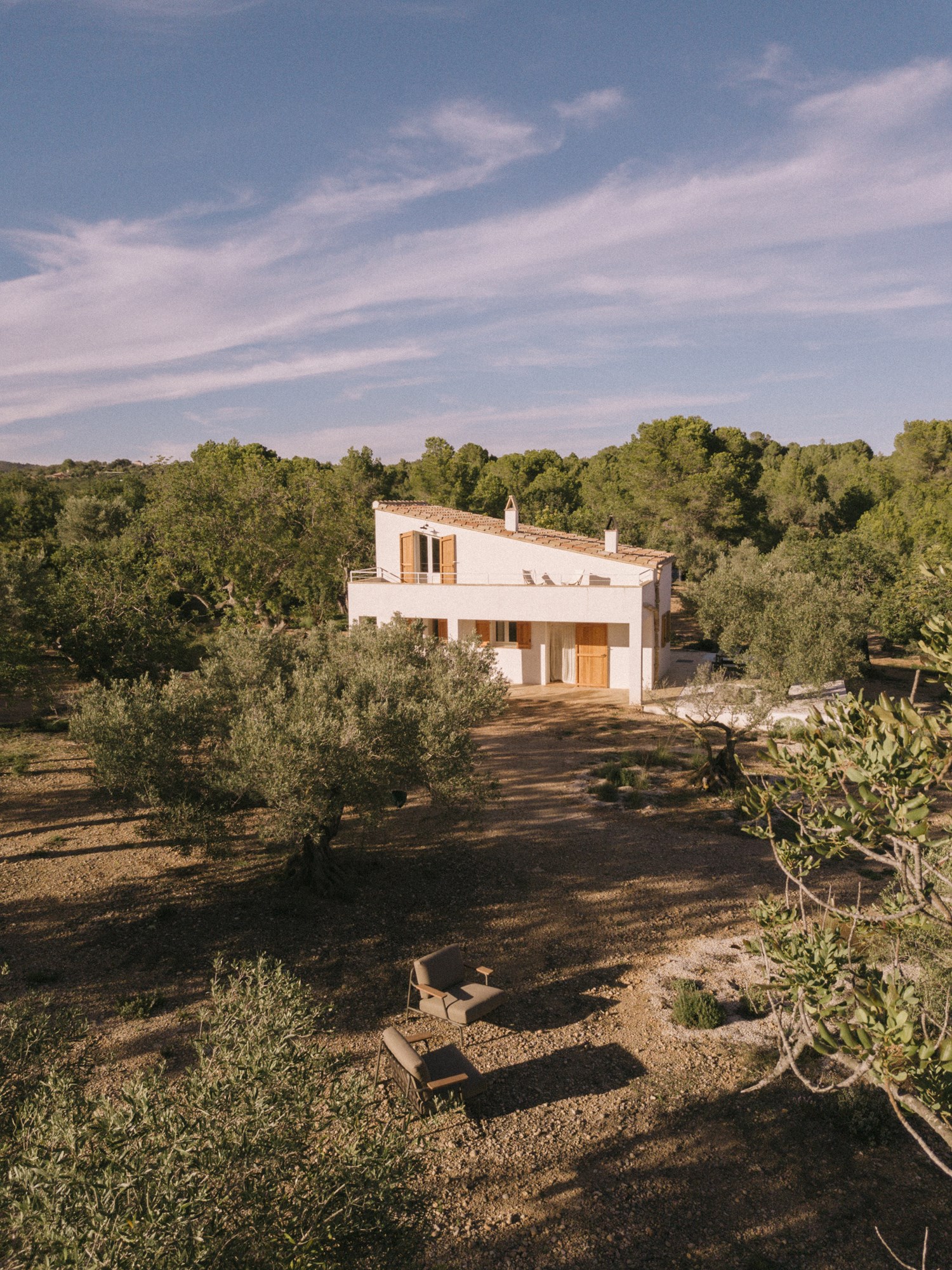 Aerial view of a Mediterranean home – A two-story white stucco house with a terracotta-tiled roof sits among olive trees, bathed in golden light. Wooden shutters and doors accentuate its rustic charm. In the foreground, a cozy outdoor seating area with two brown cushioned chairs and a bench blends seamlessly into the dry, earthy landscape. Aerial view of a Mediterranean home – A two-story white stucco house with a terracotta-tiled roof sits among olive trees, bathed in golden light. Wooden shutters and doors accentuate its rustic charm. In the foreground, a cozy outdoor seating area with two brown cushioned chairs and a bench blends seamlessly into the dry, earthy landscape.