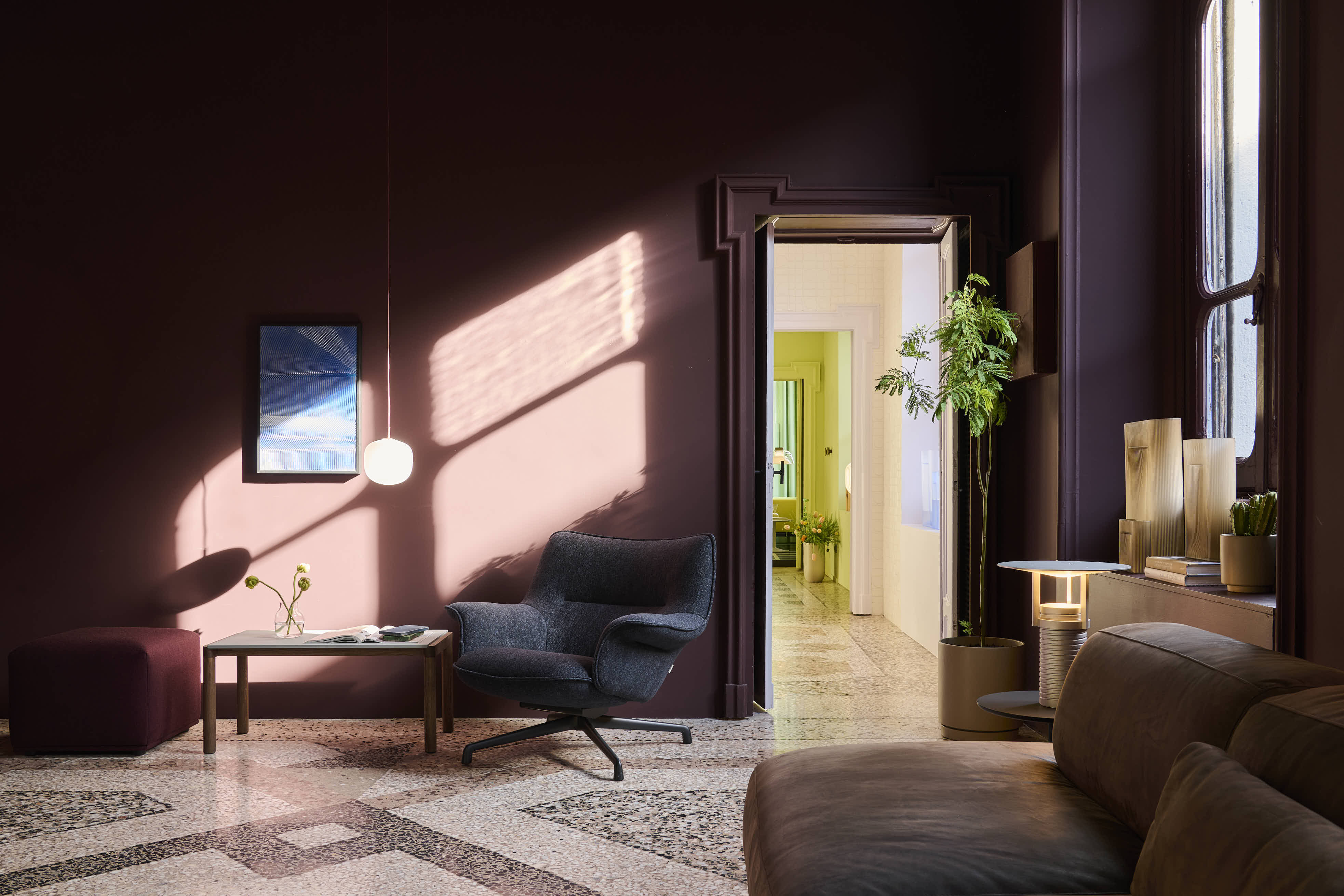 A dark room with rays of sunlight. A soft lounge chair is placed in the room, together with a sofa table, a couch, a pouf, and a pendant lamp. The room is styled with vases of different heights and a green plant.