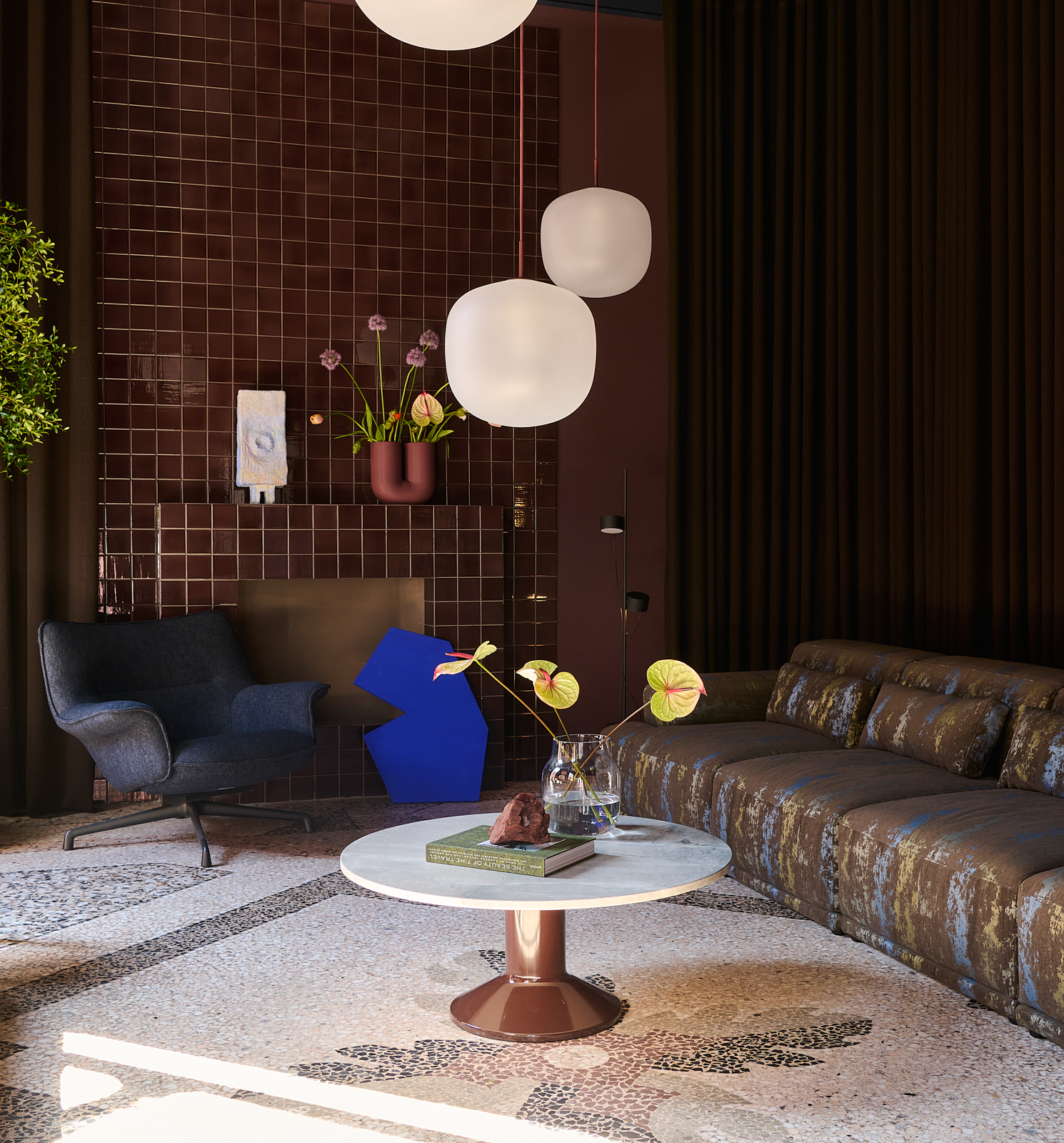 A dark blue lounge chair, a dark sofa, and a marble chair are placed in a dark burgundy room with a mosaic floor. The room is styled with two pendant lamps and artistic vases with flowers.