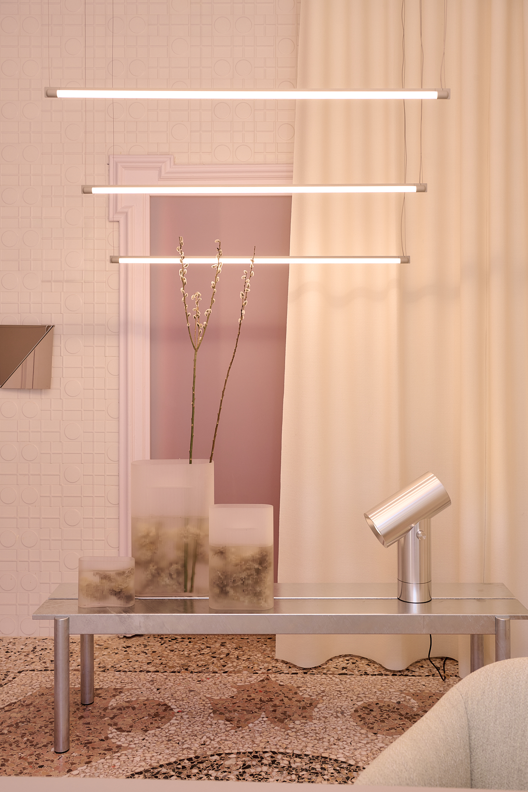 A light, bright dusty pink room styled with a metal bench and a table lamp in polished aluminium. Three vases in clear frosted glass are placed beside the lamp. Modern tube-shaped pendant lamps hang from the ceiling.