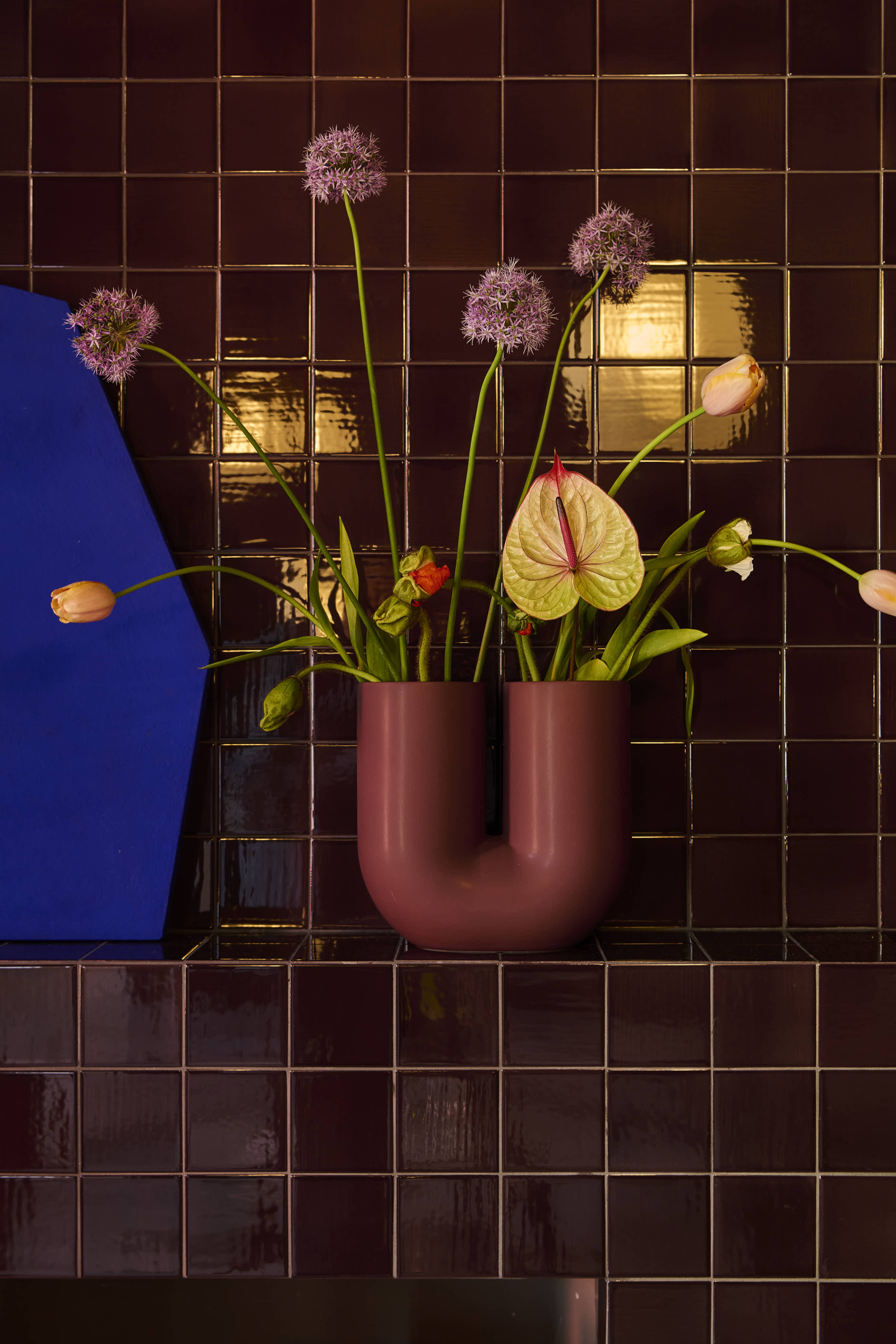 A close-up of a sculptural burgundy vase with colorful flowers, placed on a glazed stone shelf.
