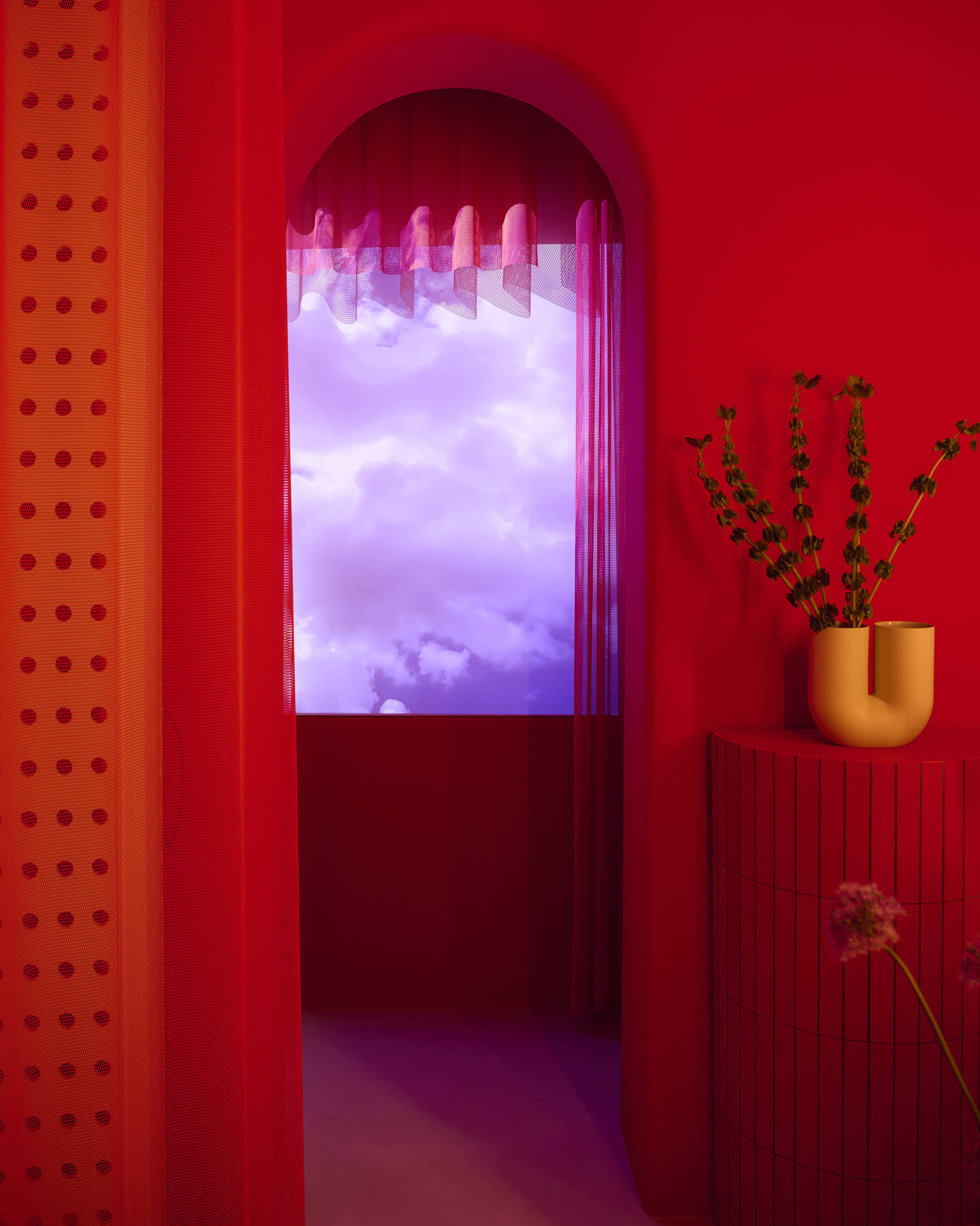 A red room with orange undertones. A pedestal is placed in the room, accompanied by a sculptural vase with flowers. Modern art showcasing a neon sky in light purple is placed beside the pedestal to create a window-like effect.