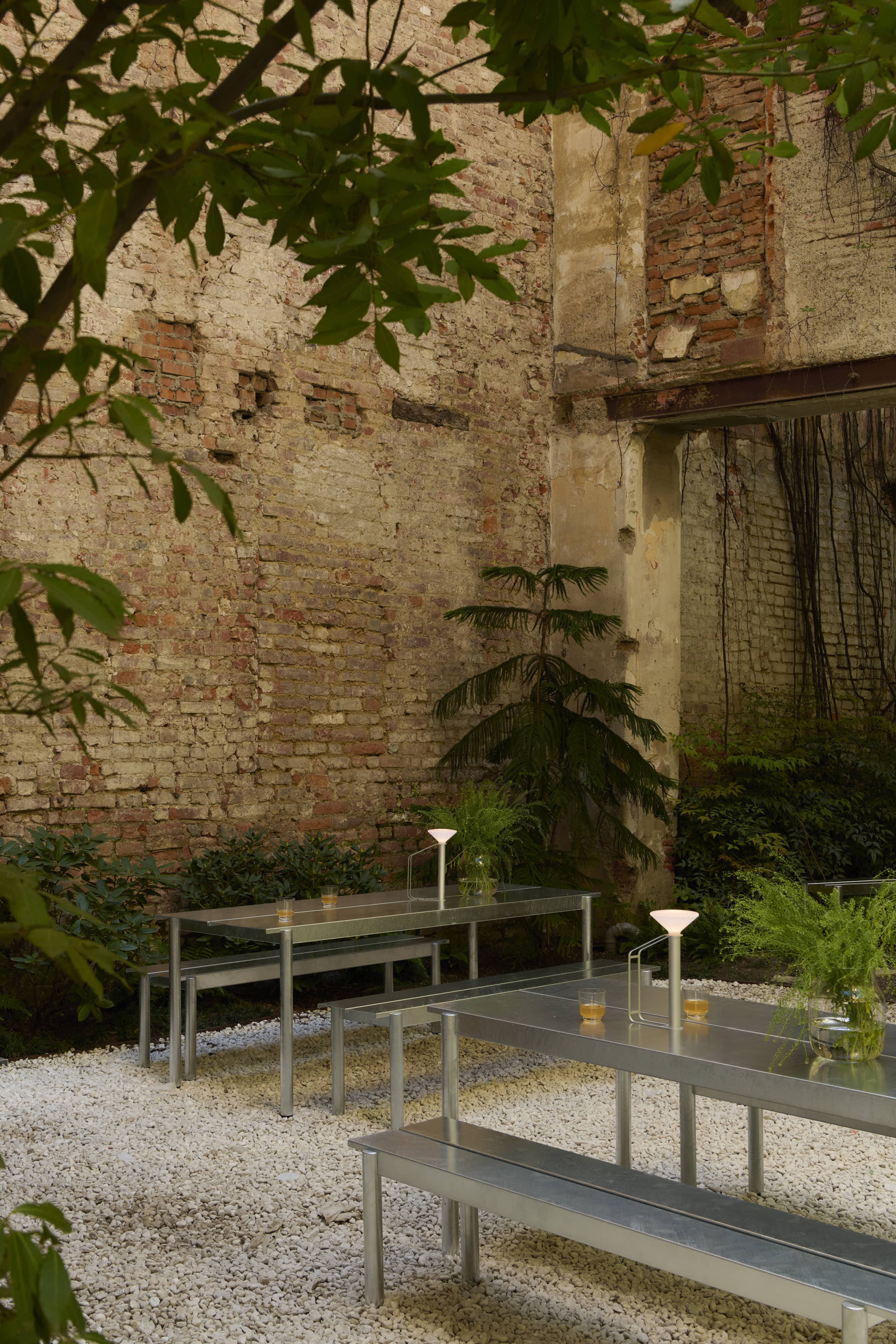 Outdoor tables and benches in polished steel are placed in a cozy courtyard. Green plants and trees are planted along the stone wall. The outdoor furniture is styled with outdoor lamps and vases containing green plants.