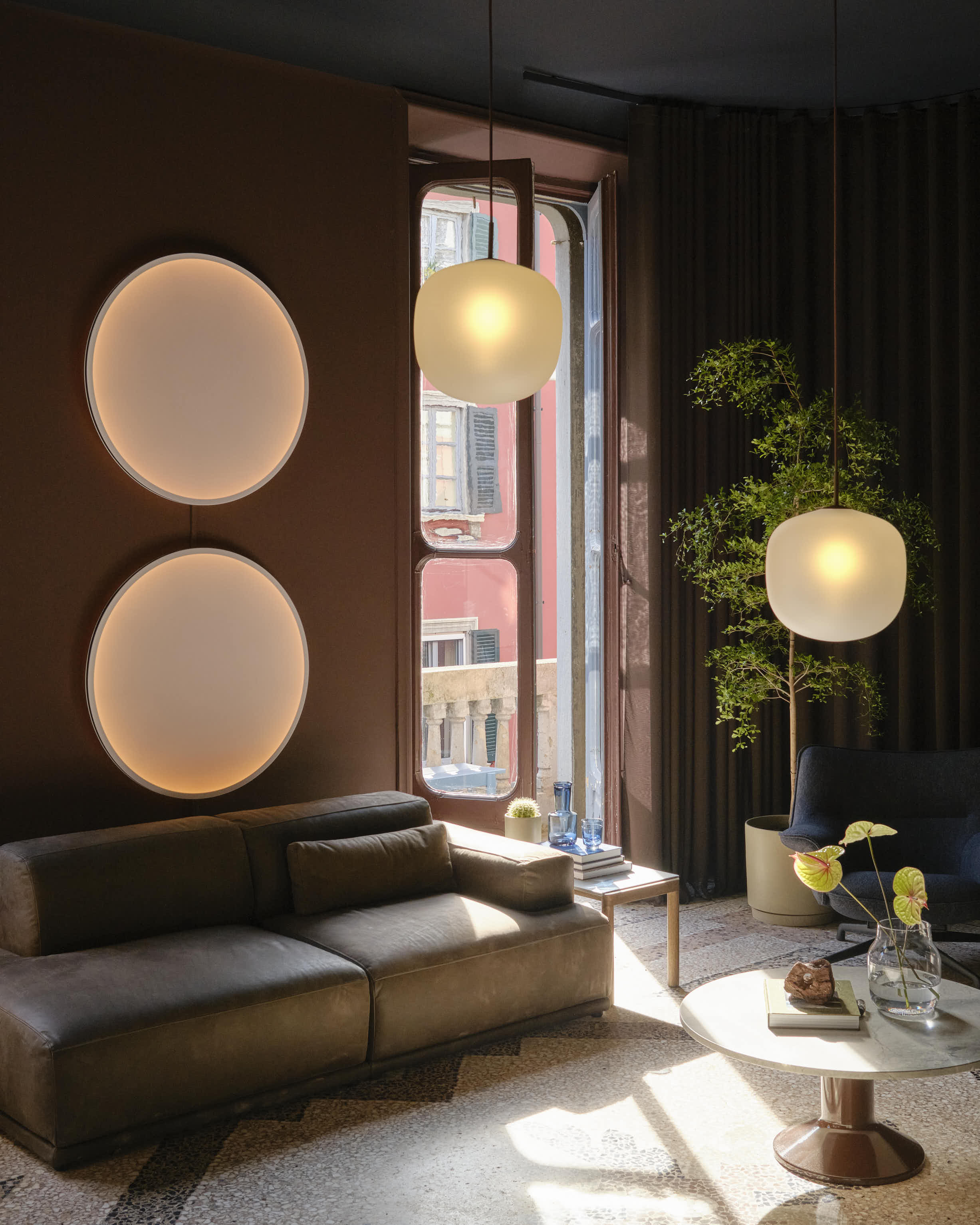 A dark burgundy room with a mosaic floor. A dark couch is placed in the room along with a marble coffee table. Two round wall lamps hang above the couch, and a pendant lamp hangs above the coffee table. A green plant is placed in the corner.