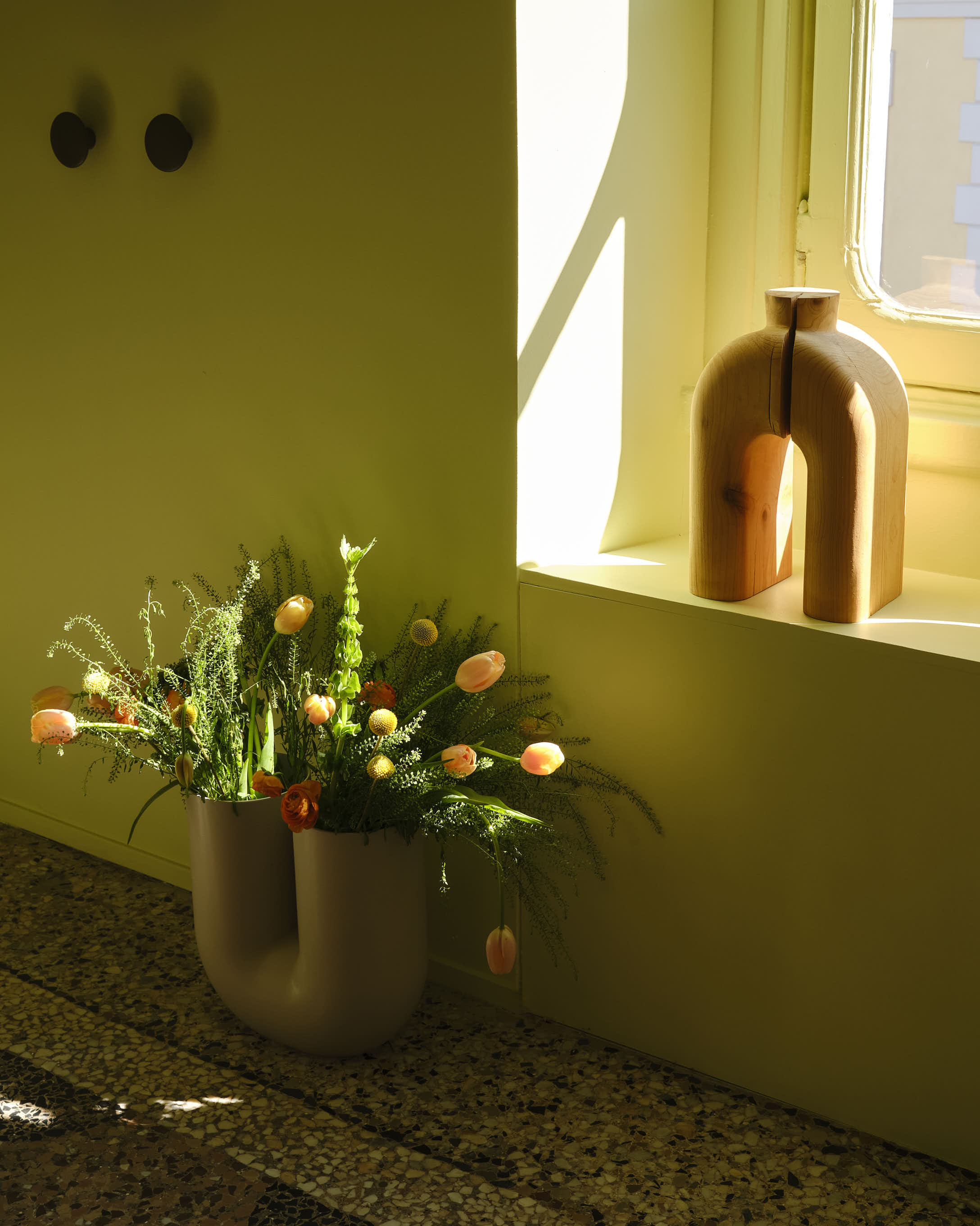 A close-up of a sculptural vase with colorful flowers placed on a mosaic floor. The wall is painted a light green, and a wooden object is placed in the window.