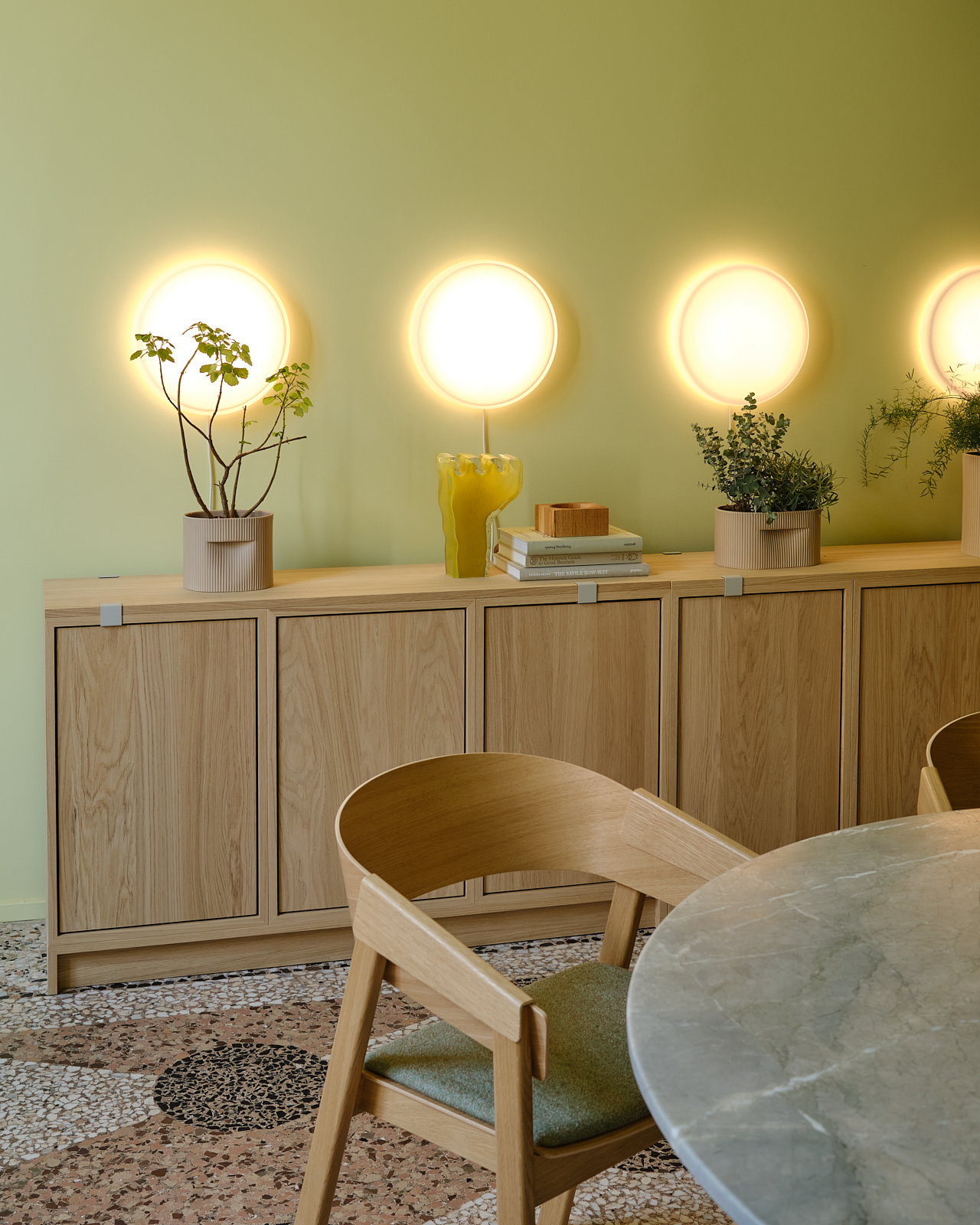 A room with a decorative mosaic floor and light green walls. Four wall lamps are aligned in a row above closed wooden sideboards. The sideboards are styled with vases and plants. A marble table is placed in the foreground, accompanied by modern wooden chairs.
