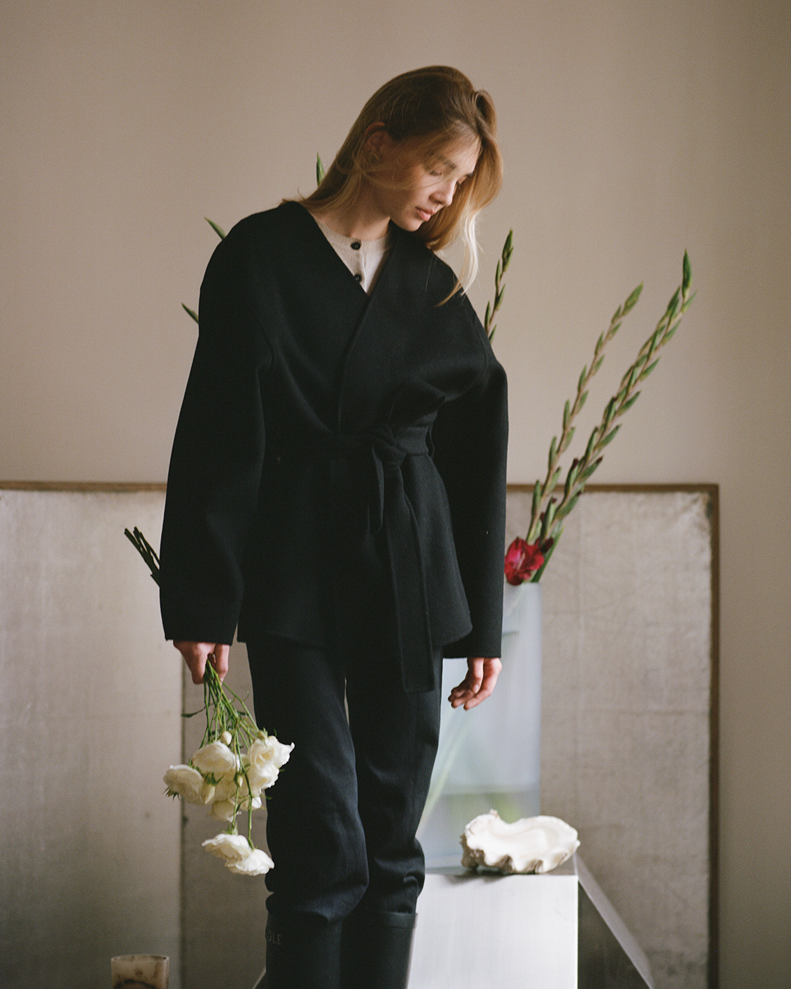 Nina stands holding a bouquet of white flowers in a black oversized jacket. Behind her, a glass vase with tall stems of greenery is displayed against a soft, neutral backdrop.
