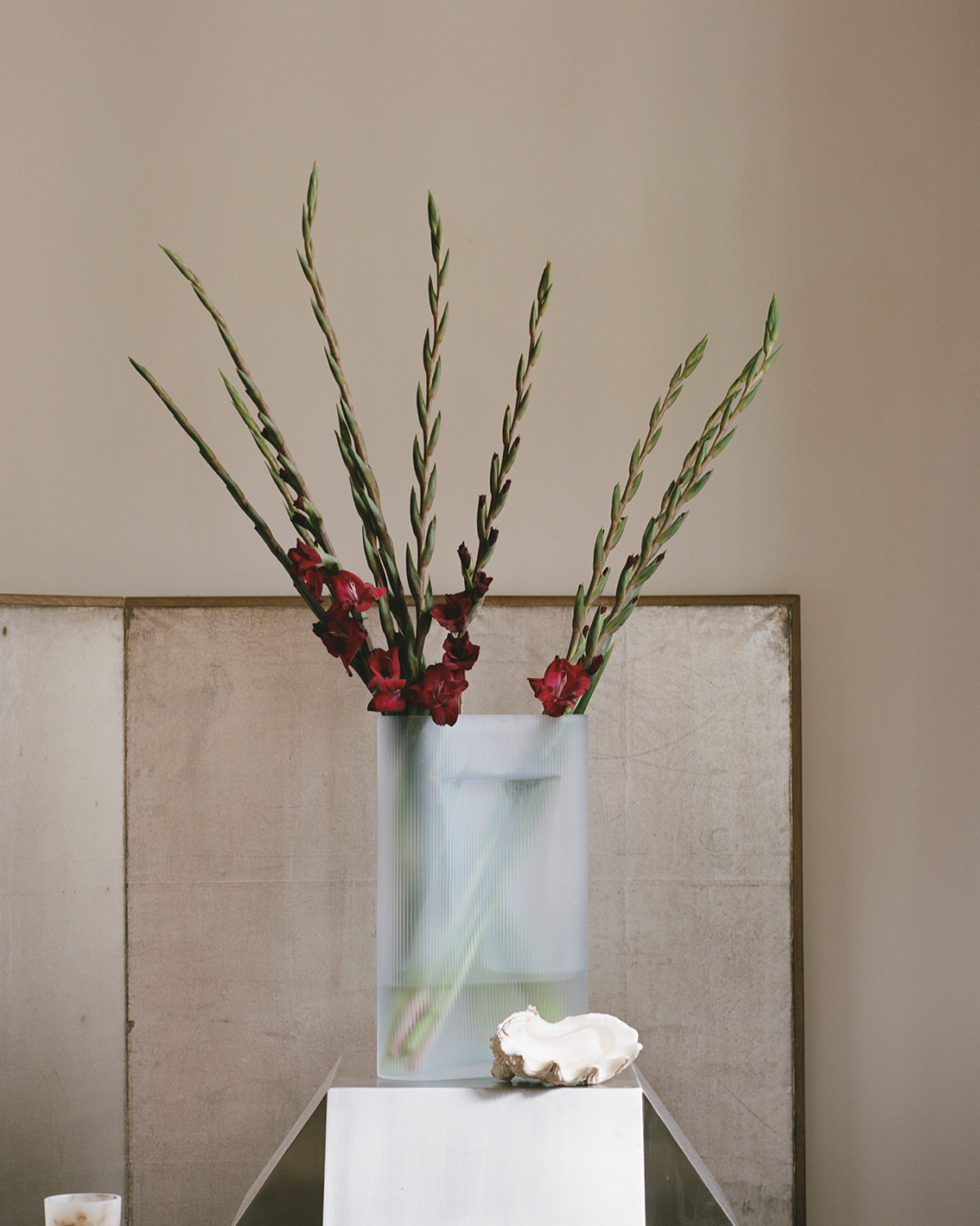 A clear glass vase with tall, slender stalks of red gladiolus flowers is placed on a minimalist pedestal. The soft, neutral background highlights the simple elegance of the floral arrangement.