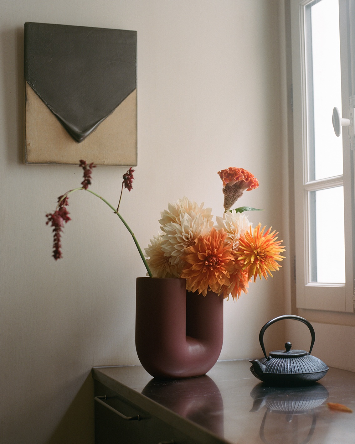A depp red U-shaped vase holds a bouquet of orange and yellow flowers, with a teapot and artwork in the background. A delicate flower stem rests against the vase.