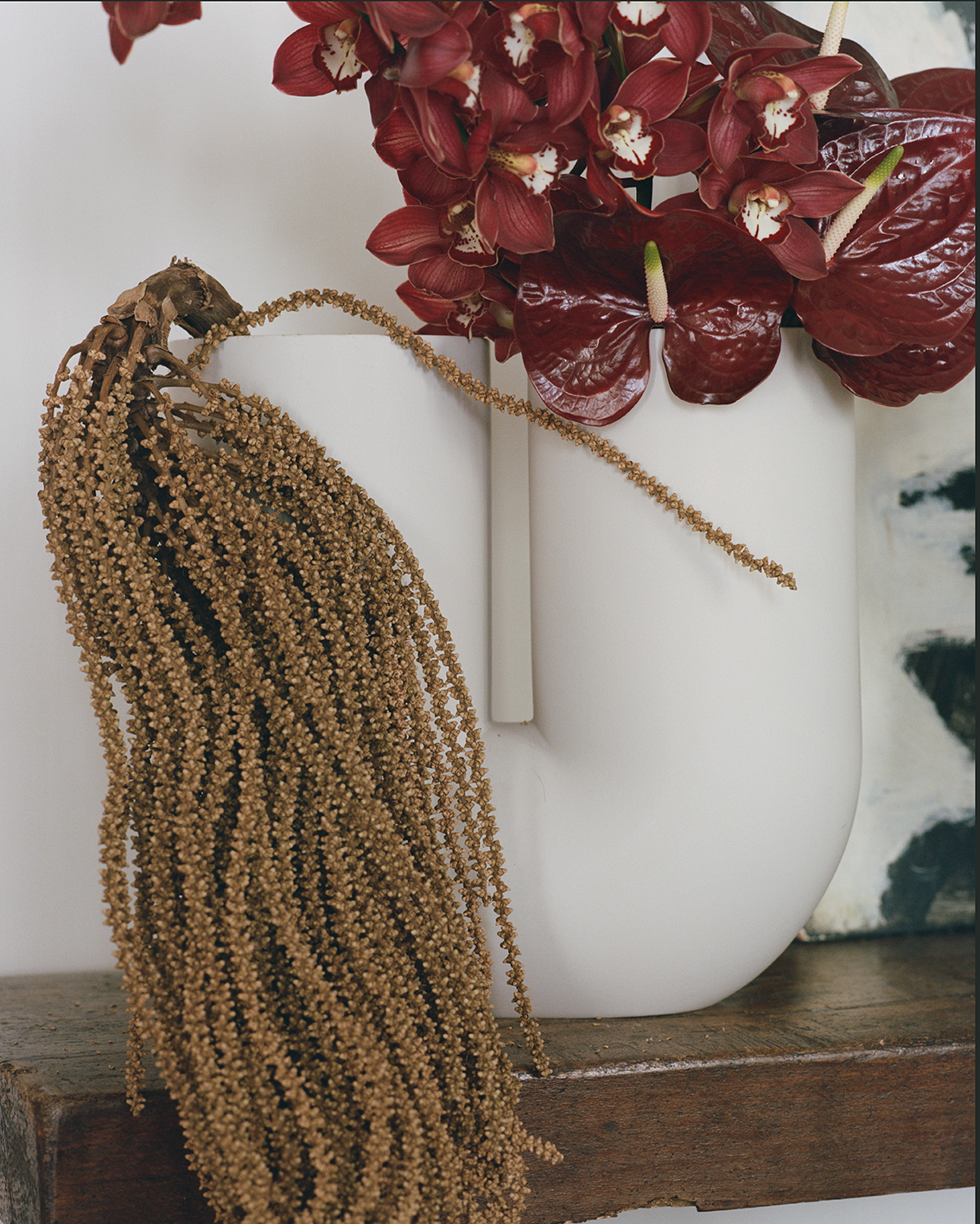 A white sculptural vase with dark red flowers and glossy red anthurium flowers is paired with dried grasses. 