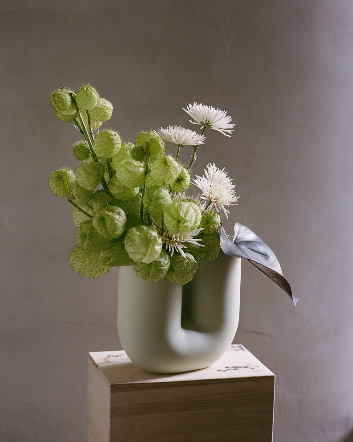 A pale U shaped green vase holds a mixture of textured green balls and white chrysanthemums. The vase is placed on a wooden pedestal in a softly lit space, highlighting the gentle tones of the flowers.