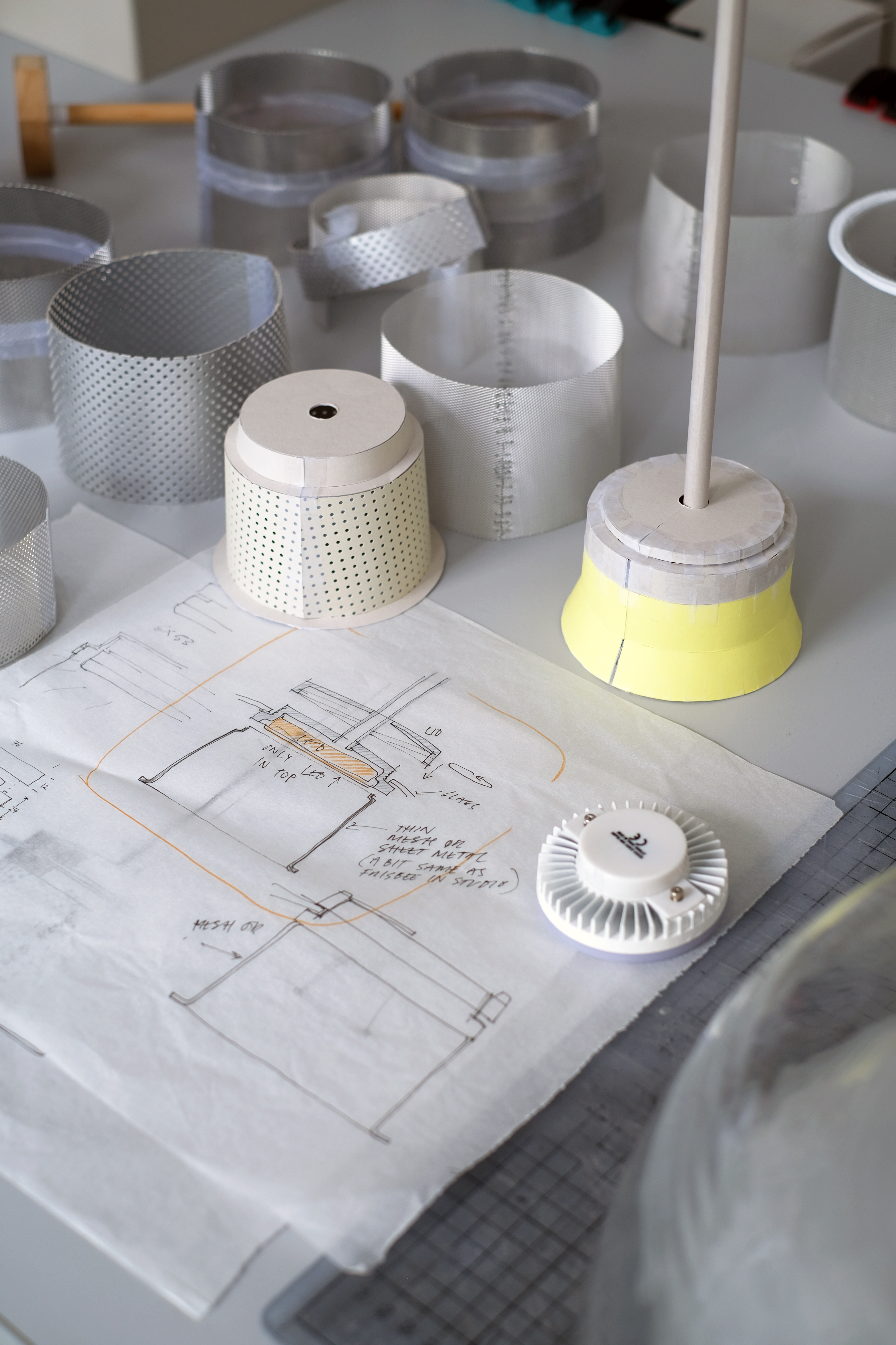 A cluttered workbench with various glass pieces, including clear bowls and metal parts, alongside paper templates. Tools and other materials are arranged across the workspace.