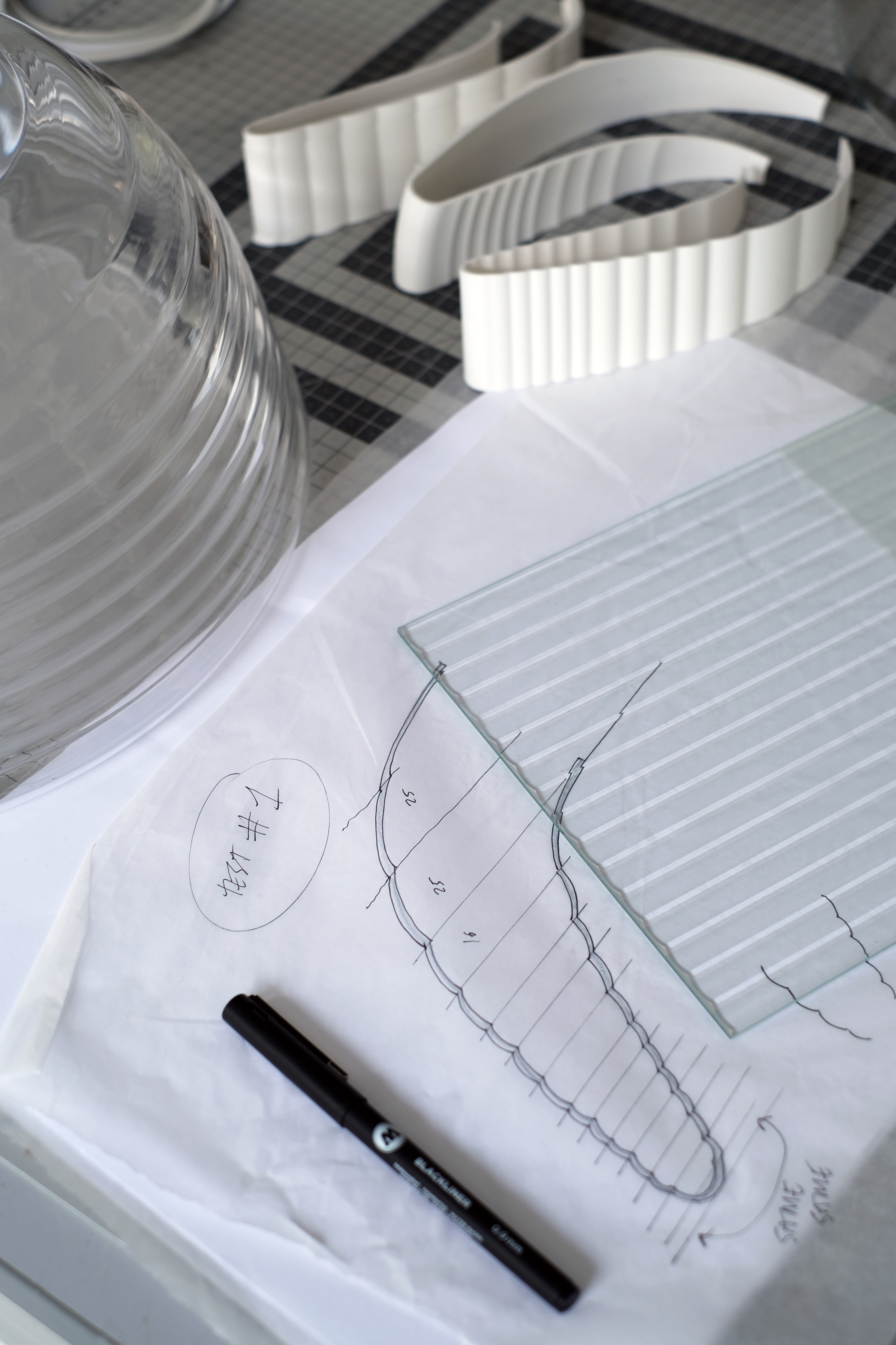 A close-up shot of a piece of glass with a ribbed pattern, with sketches on paper showing the design process. A black marker is visible in the foreground.