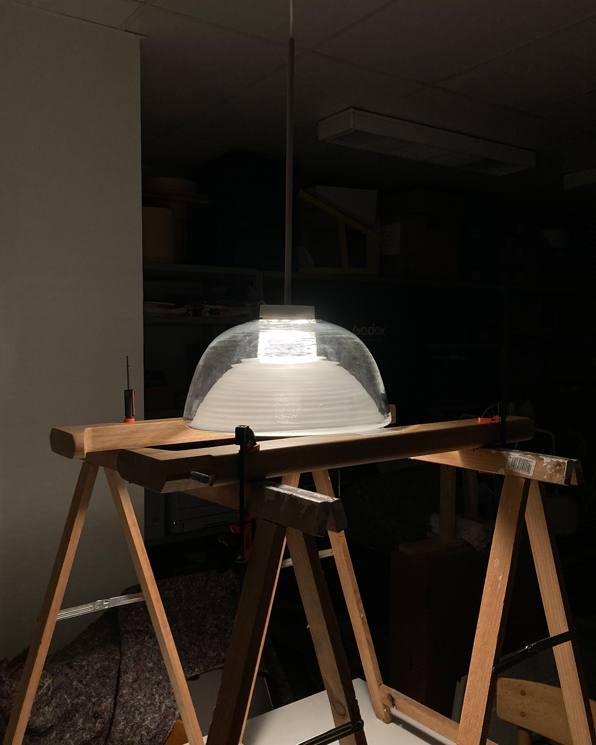 A pendant lamp with a ribbed glass shade is lit up and placed on a wooden sawhorse. The lamp's light is the primary source of illumination, casting shadows on the surrounding dark room.