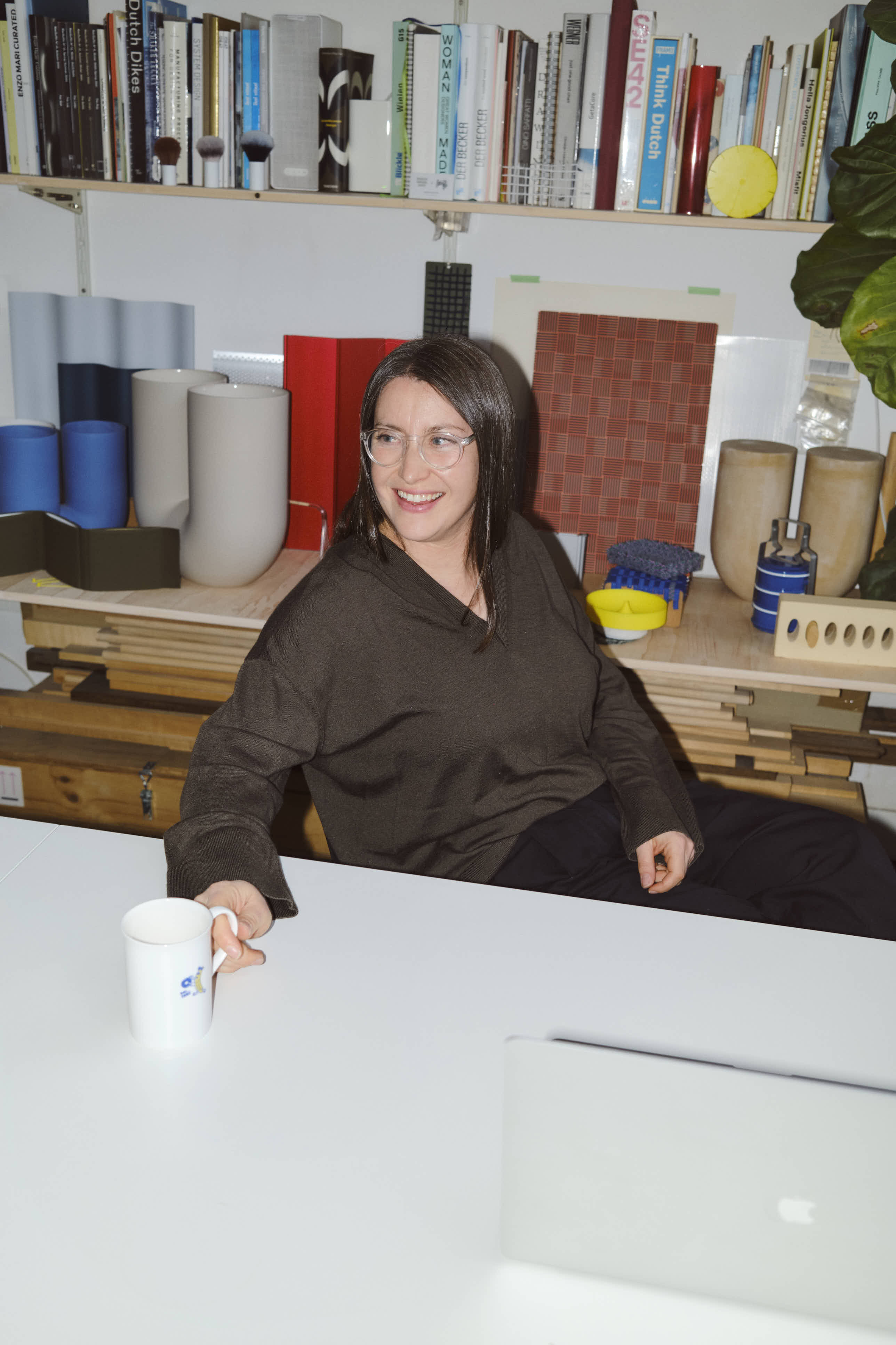 Rachel Griffin smiles while seated at a white desk in her studio, holding a white ceramic mug. Behind her, several Kink Vases in matte blue and neutral tones are displayed among material samples, tools, and a large collection of design books.