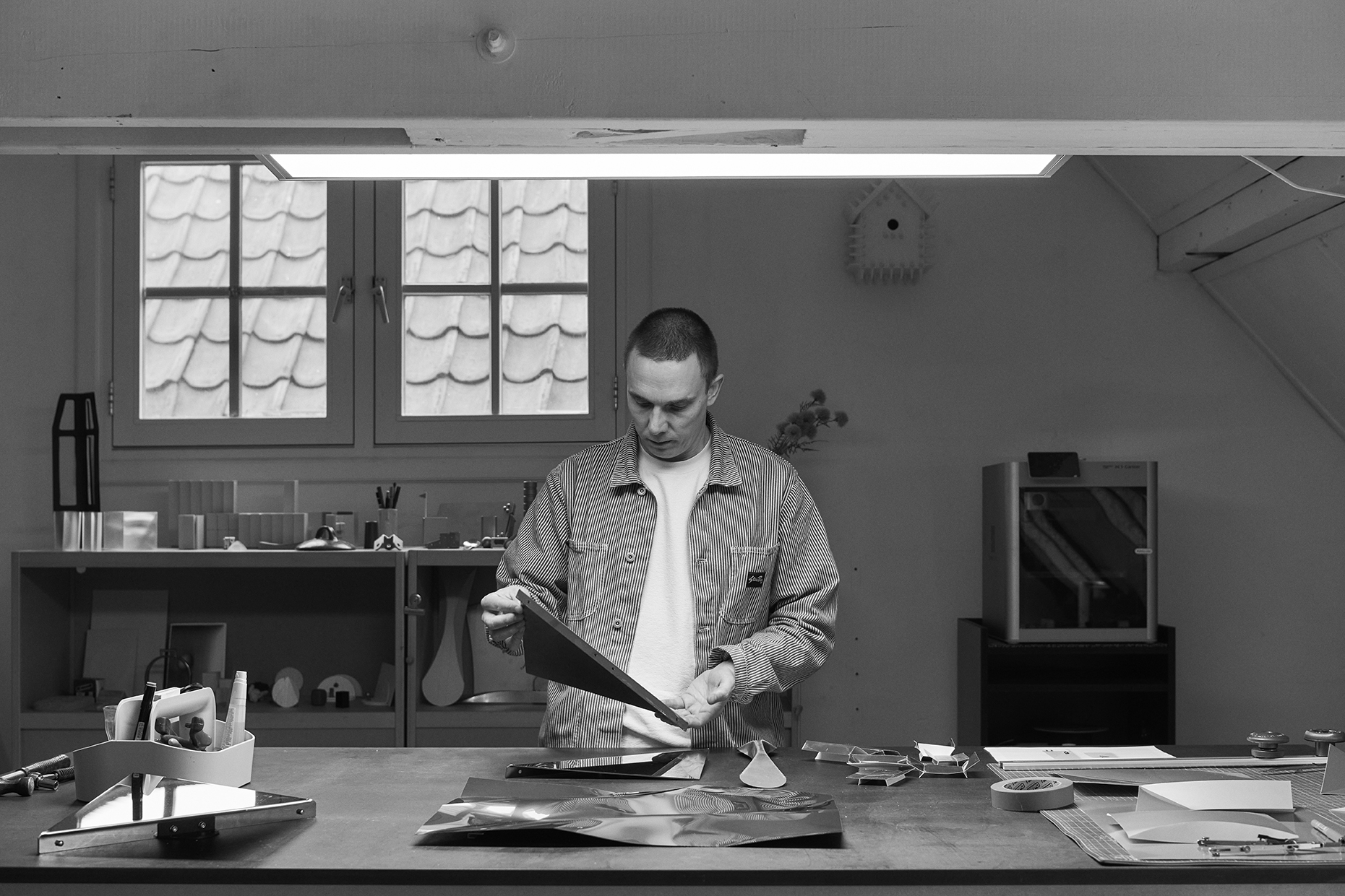 Phil in a striped jacket stands at a large worktable in a modern design studio, carefully examining a sheet of folded reflective metal.