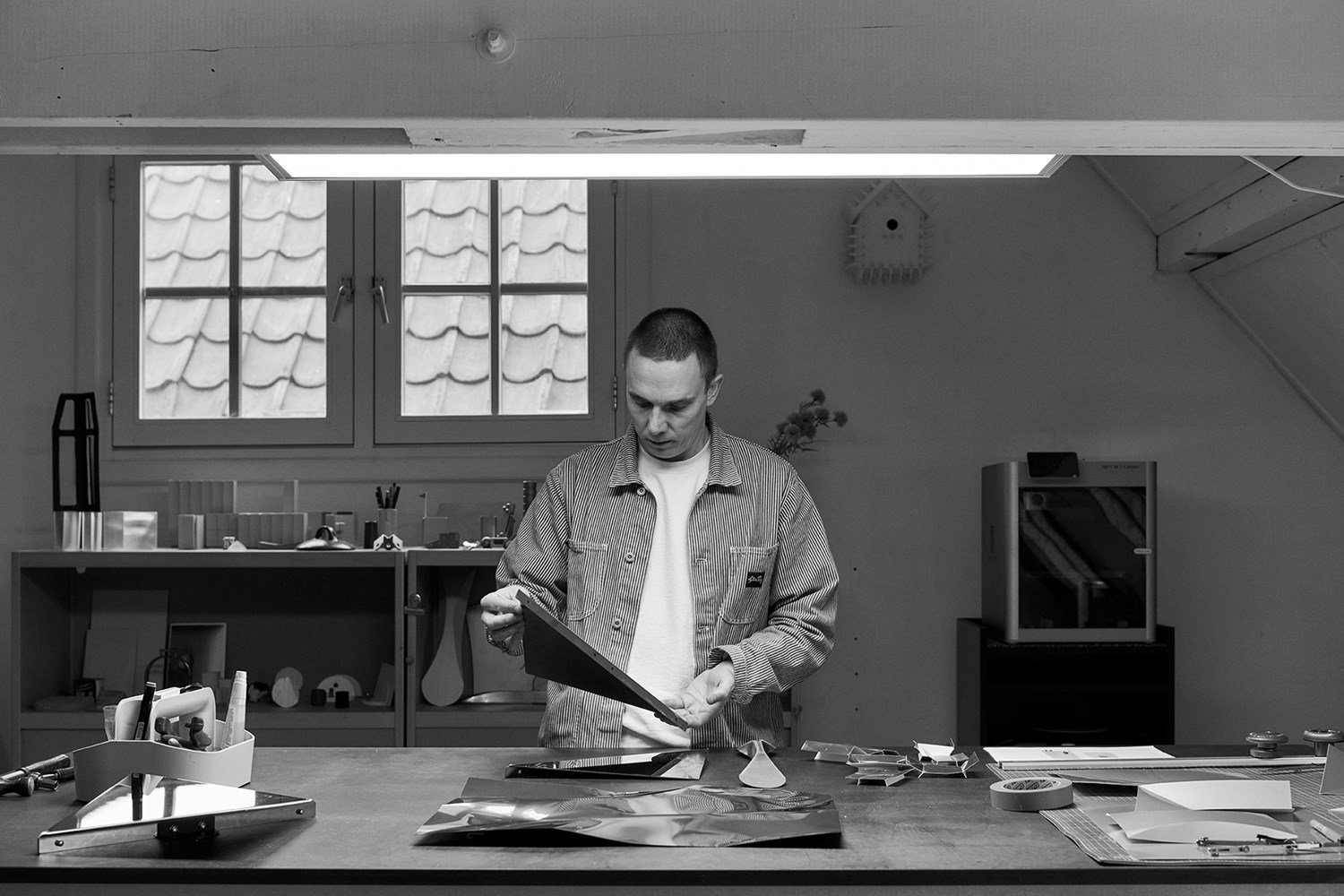 Phil in a striped jacket stands at a large worktable in a modern design studio, carefully examining a sheet of folded reflective metal. Phil in a striped jacket stands at a large worktable in a modern design studio, carefully examining a sheet of folded reflective metal.