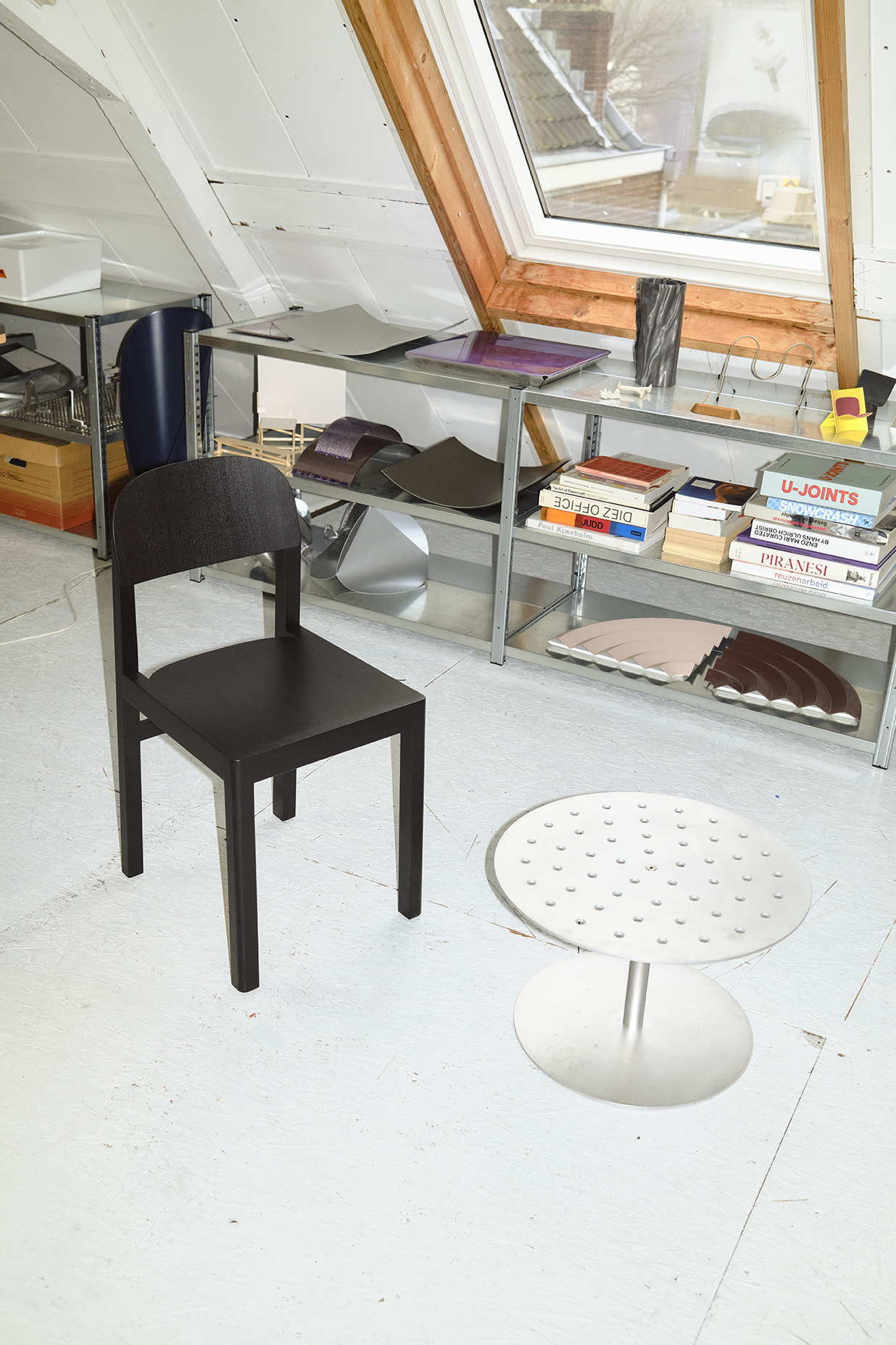 A black minimalist chair with straight legs sits beside a circular perforated metal side table in a bright attic workspace. Behind them, industrial shelving units hold sculptural metal samples, books, and tools beneath a large skylight framed in raw wood.
