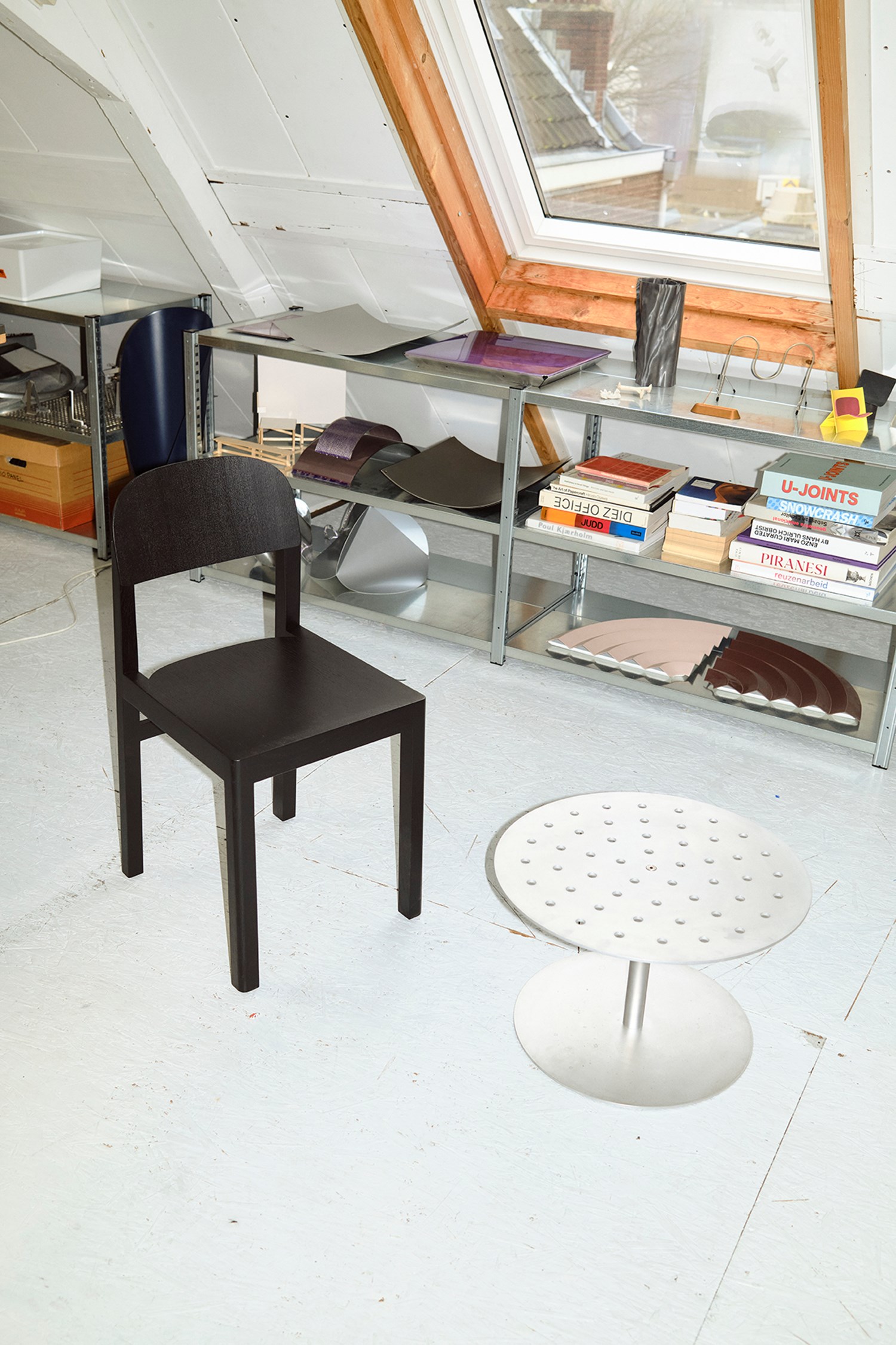 A black minimalist chair with straight legs sits beside a circular perforated metal side table in a bright attic workspace. Behind them, industrial shelving units hold sculptural metal samples, books, and tools beneath a large skylight framed in raw wood.
 A black minimalist chair with straight legs sits beside a circular perforated metal side table in a bright attic workspace. Behind them, industrial shelving units hold sculptural metal samples, books, and tools beneath a large skylight framed in raw wood.