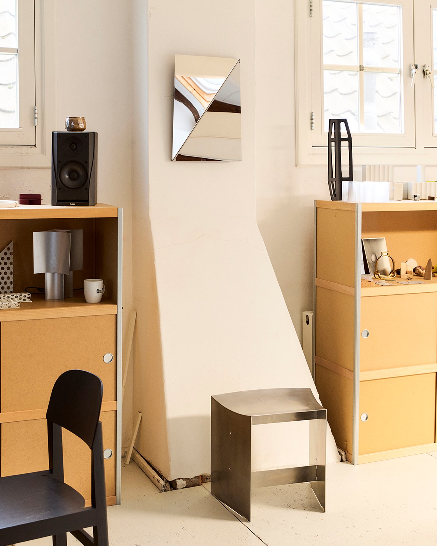 The Gaze Mirror hangs on a sloped white column between two wood and metal shelving units filled with objects and tools. Below the mirror is a minimalist, brushed steel stool with a softly curved seat.
 The Gaze Mirror hangs on a sloped white column between two wood and metal shelving units filled with objects and tools. Below the mirror is a minimalist, brushed steel stool with a softly curved seat.