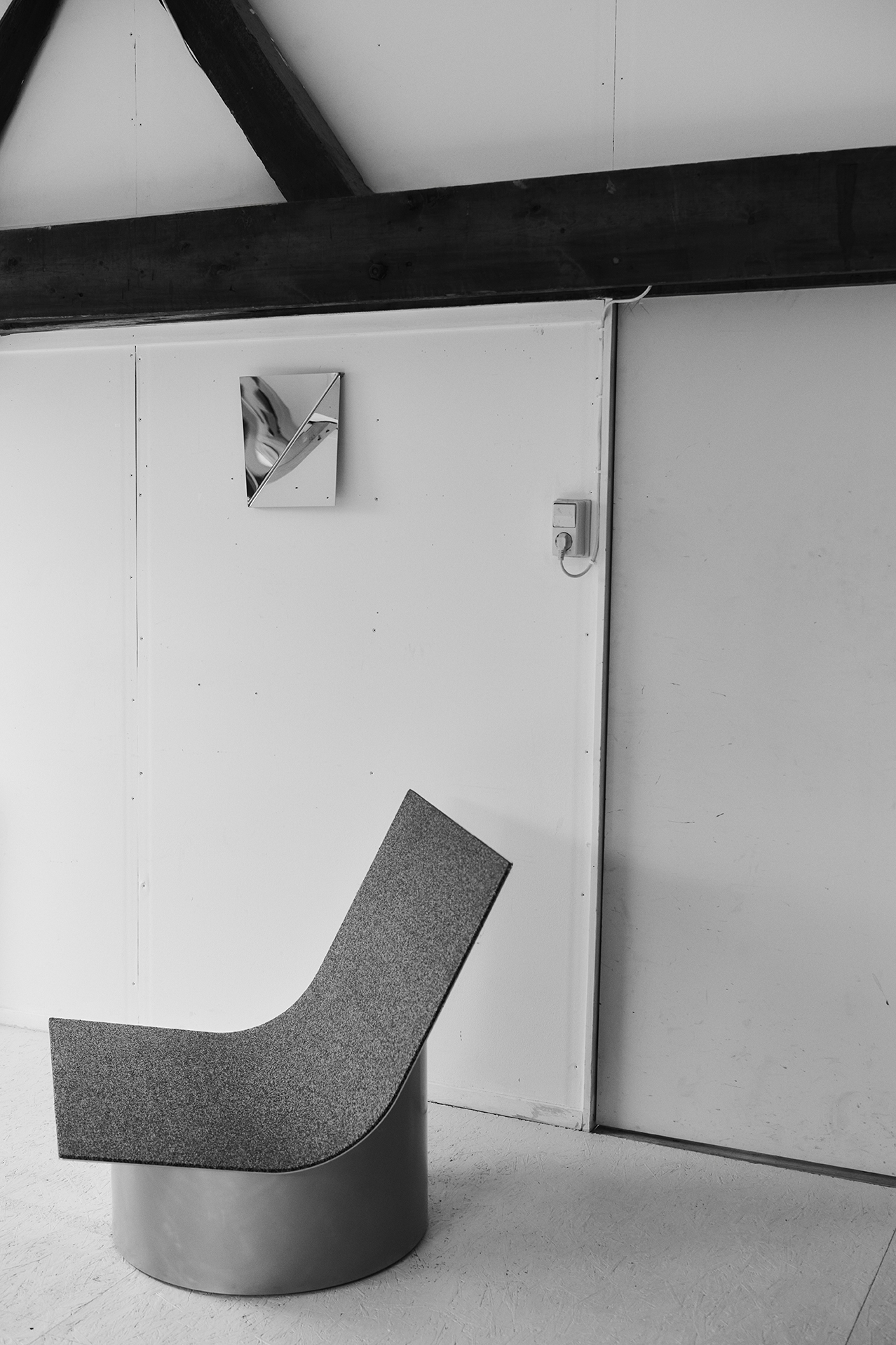 A sculptural chair with a cylindrical metal base and a high, sweeping backrest upholstered in textured fabric sits against a white wall with exposed wooden beams. Above it hangs the Gaze Mirror composed of angled polished panels, creating optical distortions.
