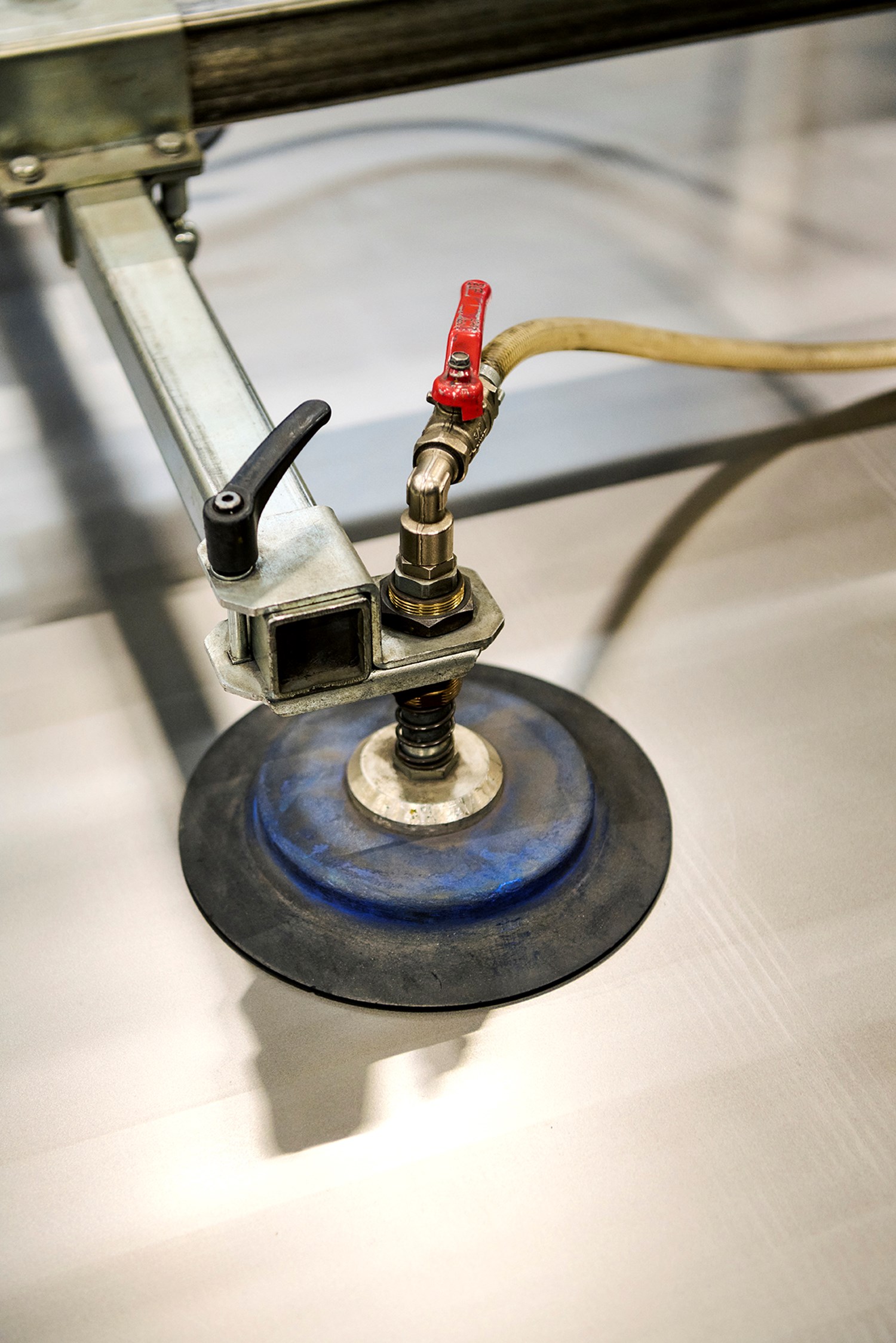 Detailed close-up of a suction-based vacuum lifting tool with metal arms, a black rubber pad, and red valve handle, positioned on a smooth metal sheet. The equipment highlights industrial material handling in a precision-focused design or fabrication environment.
 Detailed close-up of a suction-based vacuum lifting tool with metal arms, a black rubber pad, and red valve handle, positioned on a smooth metal sheet. The equipment highlights industrial material handling in a precision-focused design or fabrication environment.