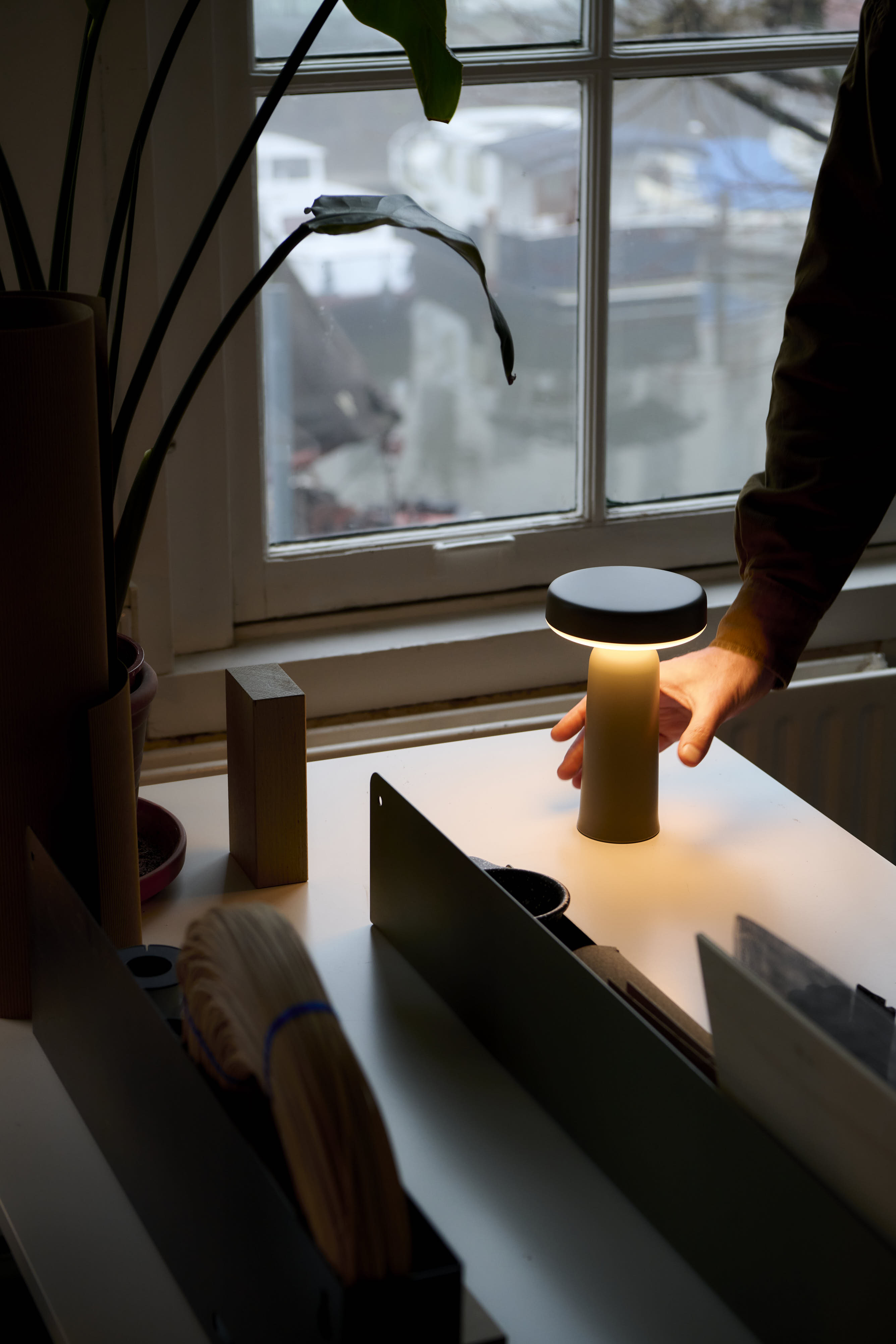 Johan’s hand reaches for a glowing Muuto Ease portable lamp placed on a desk near a window overlooking a canal, with plants and office supplies around.