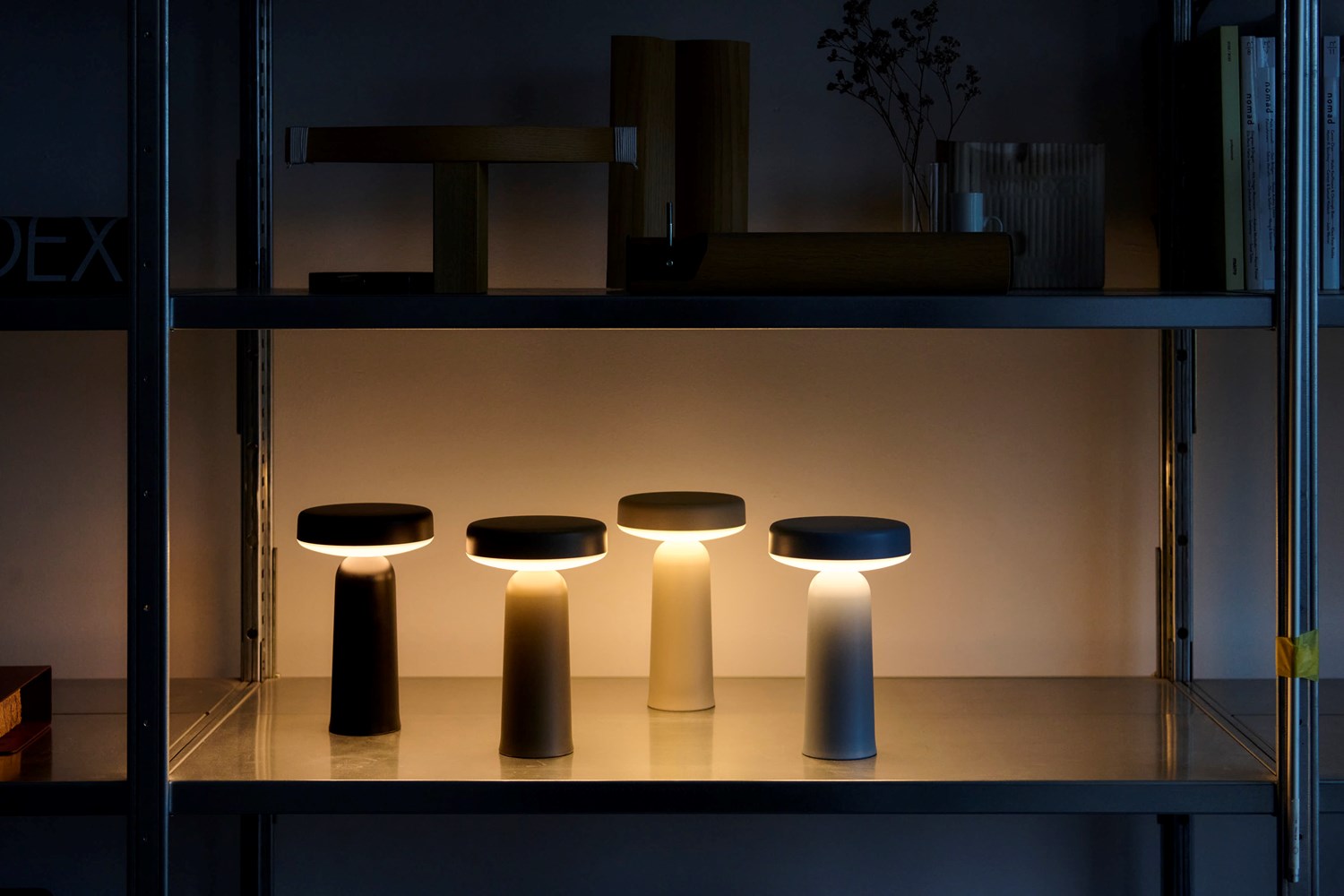 Four Muuto Ease portable lamps glowing in a dimly lit room, placed on a metal shelf among wooden and metal objects, creating a warm ambiance. Four Muuto Ease portable lamps glowing in a dimly lit room, placed on a metal shelf among wooden and metal objects, creating a warm ambiance.
