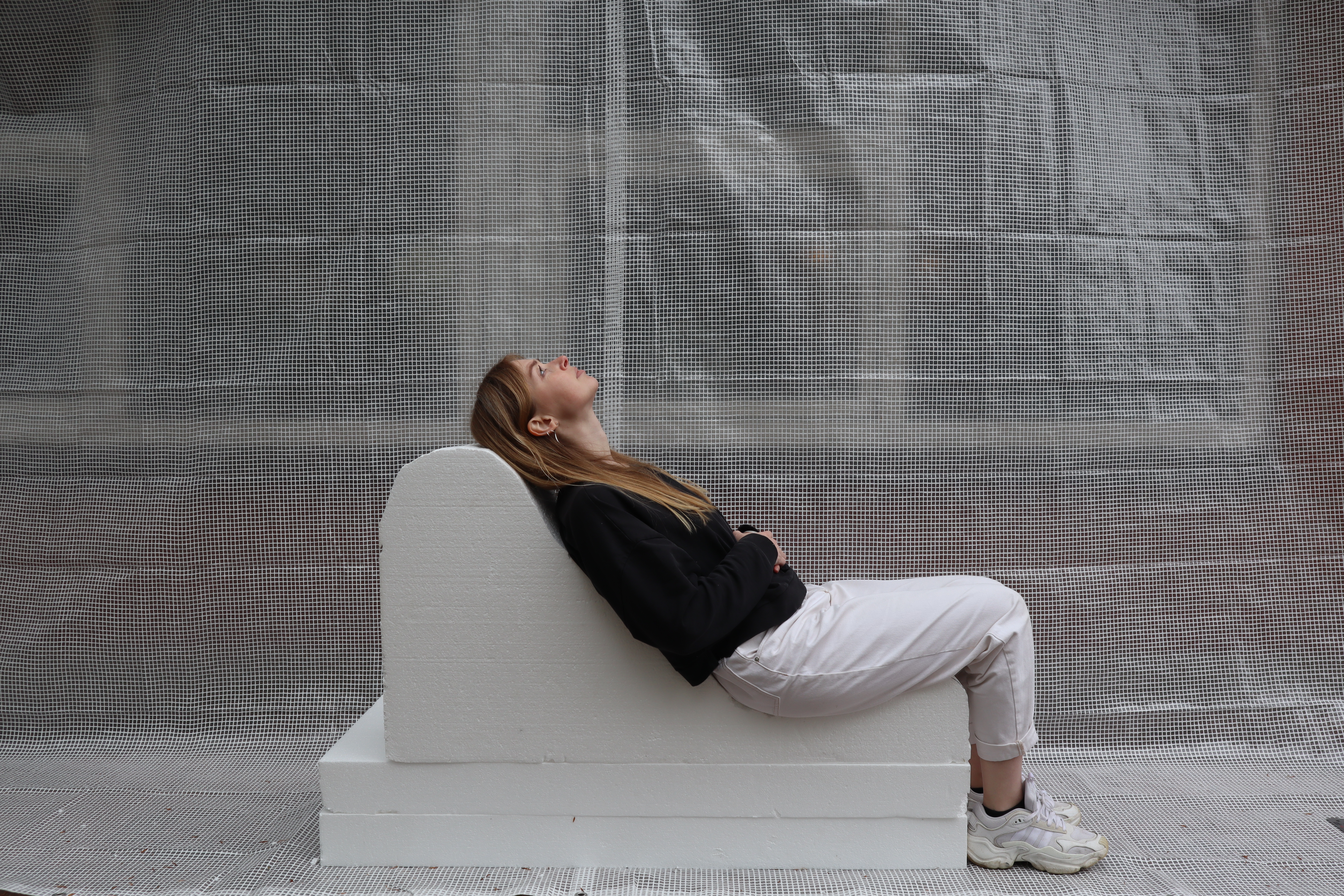 Lise Vester sitting on the Dream View Bench in a raw, industrial space with concrete flooring and minimal surroundings, creating a contrast between the soft, sculptural bench and the rough setting.