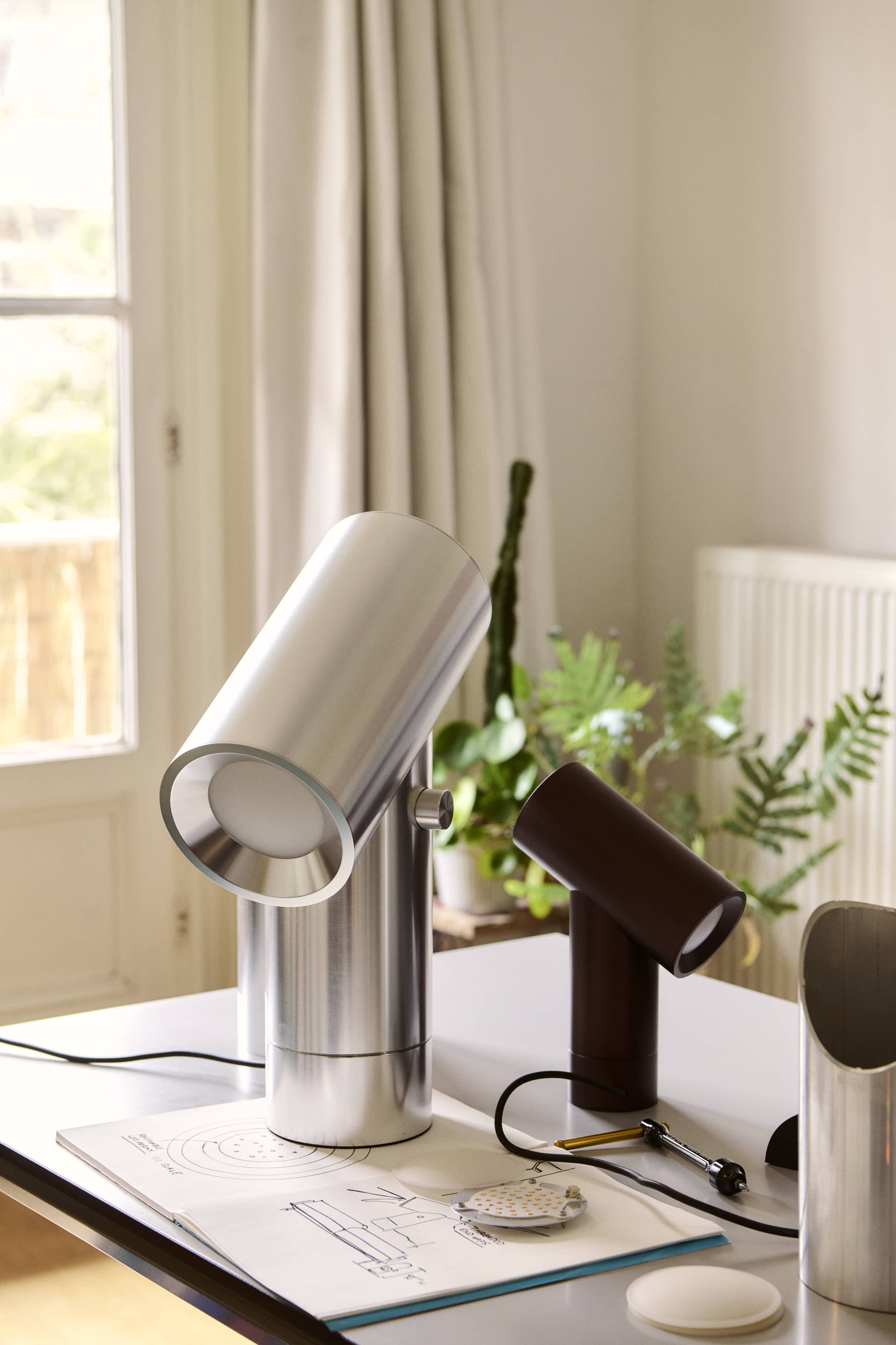 A Modern setting with silver and black Beam table lamps on a desk with papers, near a window and indoor plants.