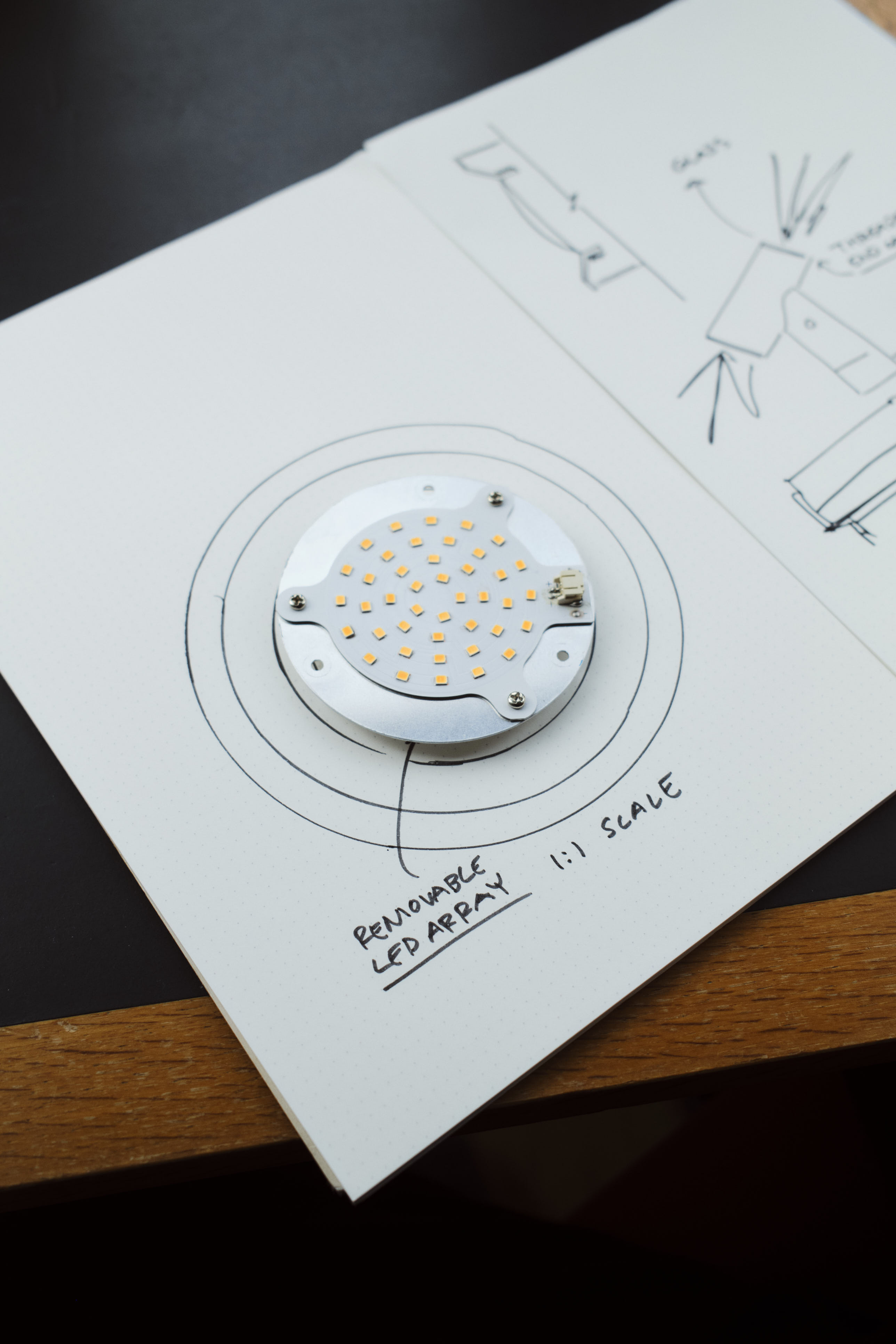 An open notebook with hand-drawn sketches and notes on lighting design rests on a dark table with wooden trim. A circular LED component sits on the page, aligning with the technical diagrams.
