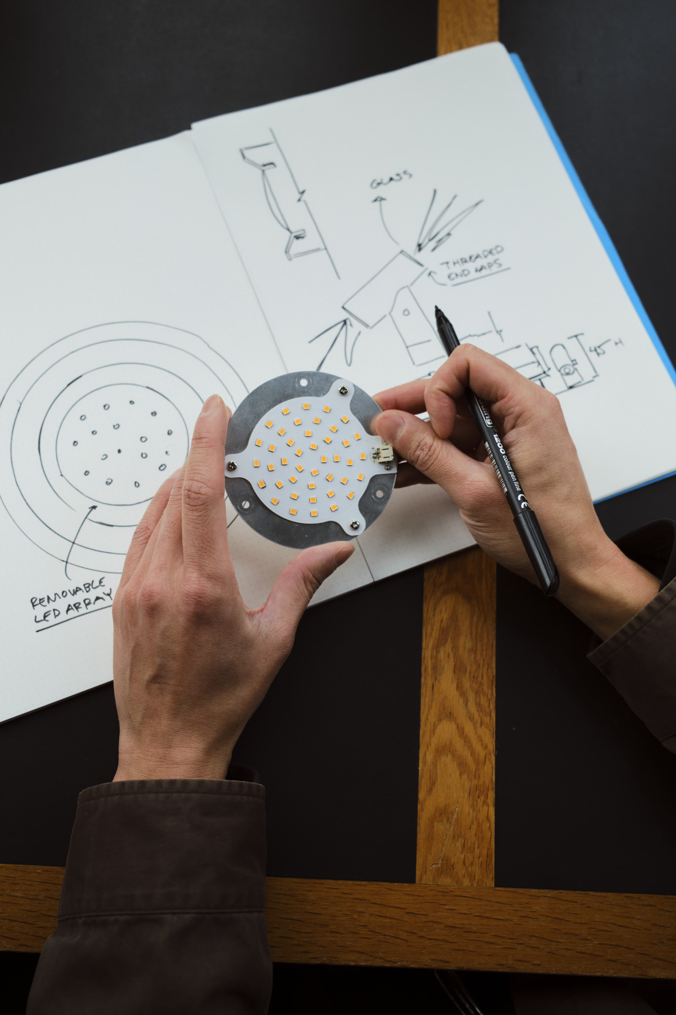 A pair of hands holds a circular LED component while sketching design ideas in a notebook. The open pages feature hand-drawn diagrams and notes. The dark table with wooden trim provides a warm, creative workspace.