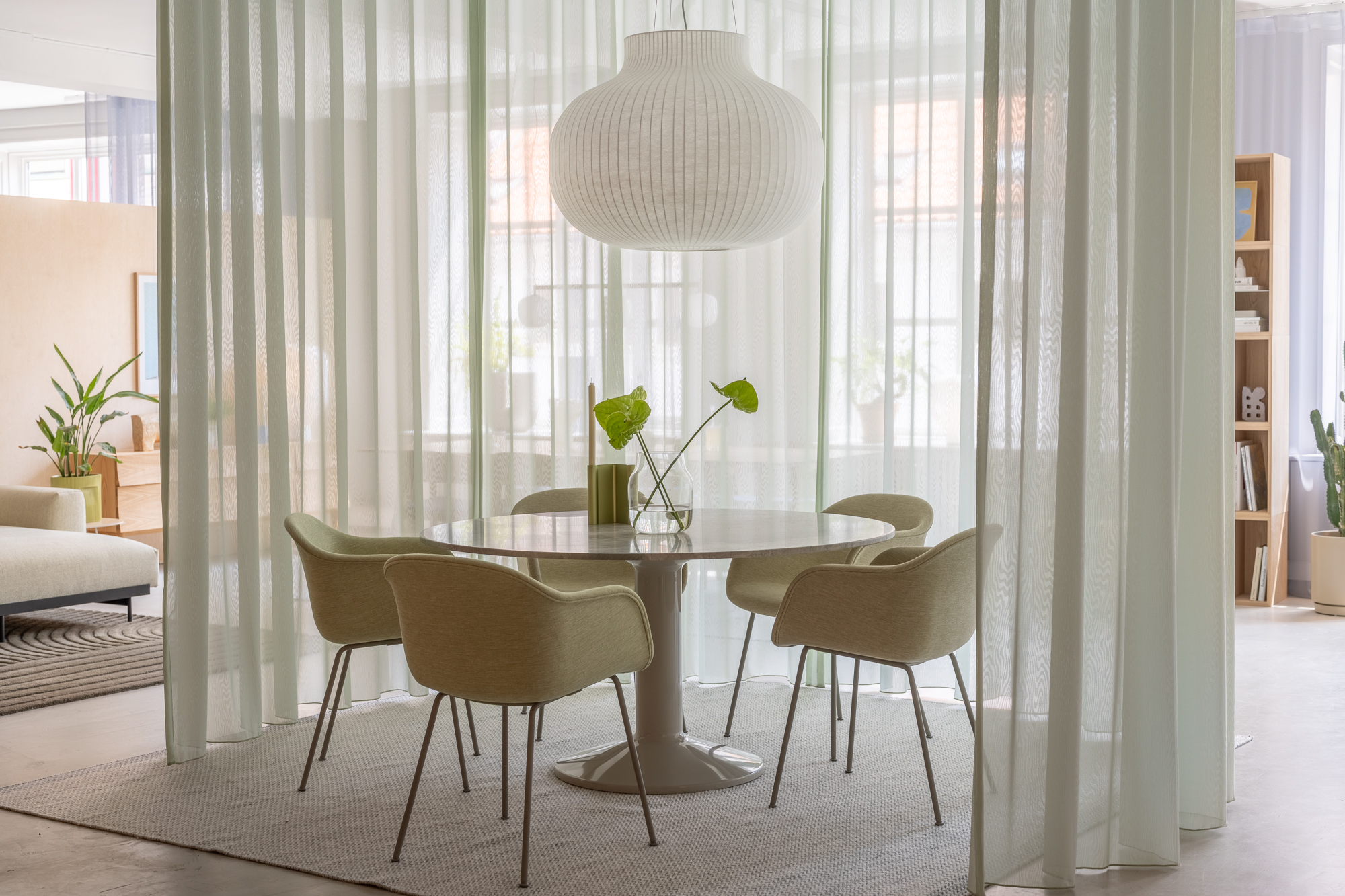 The Midst table in marble is placed in a white room with modern curtains. Soft green chairs surround the table, while a pendant lamp hangs above, adding a cozy touch to the space.