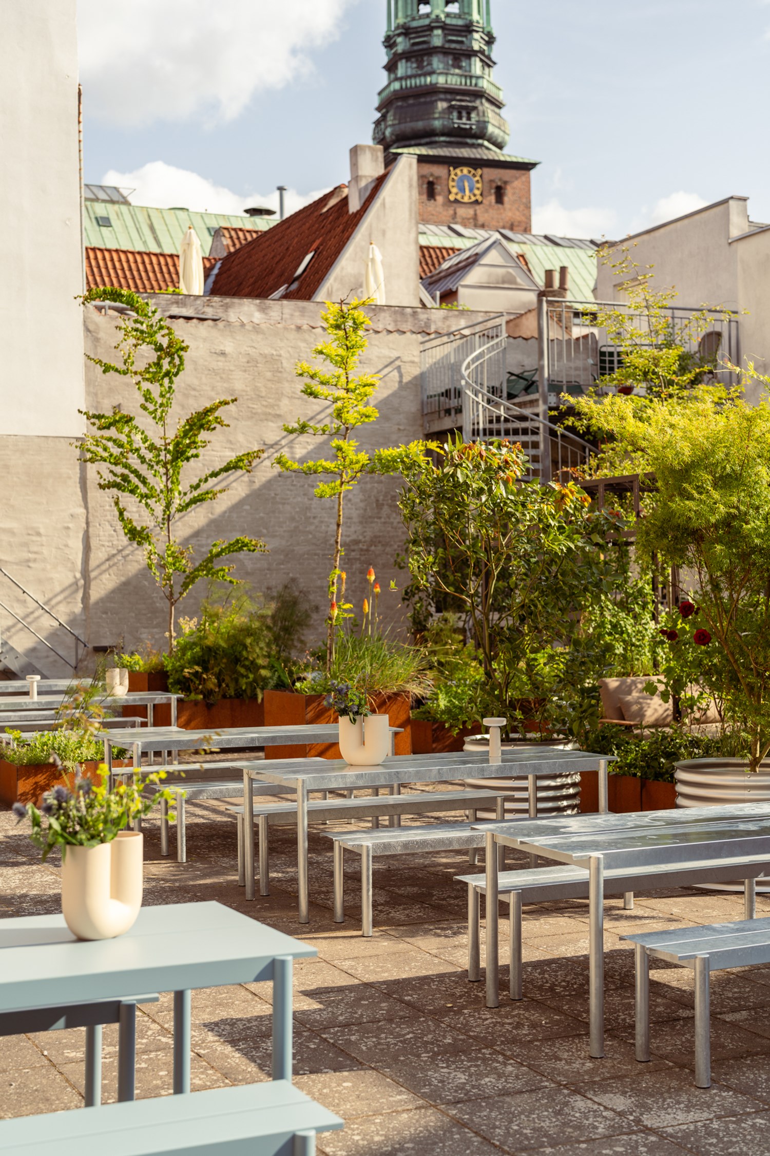 Linear Steel tables and benches in light blue are arranged in two rows in a cozy backyard surrounded by green trees and lush plants. The artistic Kink Vase sits atop the tables, styled with a creative floral arrangement that adds a sculptural touch. Linear Steel tables and benches in light blue are arranged in two rows in a cozy backyard surrounded by green trees and lush plants. The artistic Kink Vase sits atop the tables, styled with a creative floral arrangement that adds a sculptural touch.