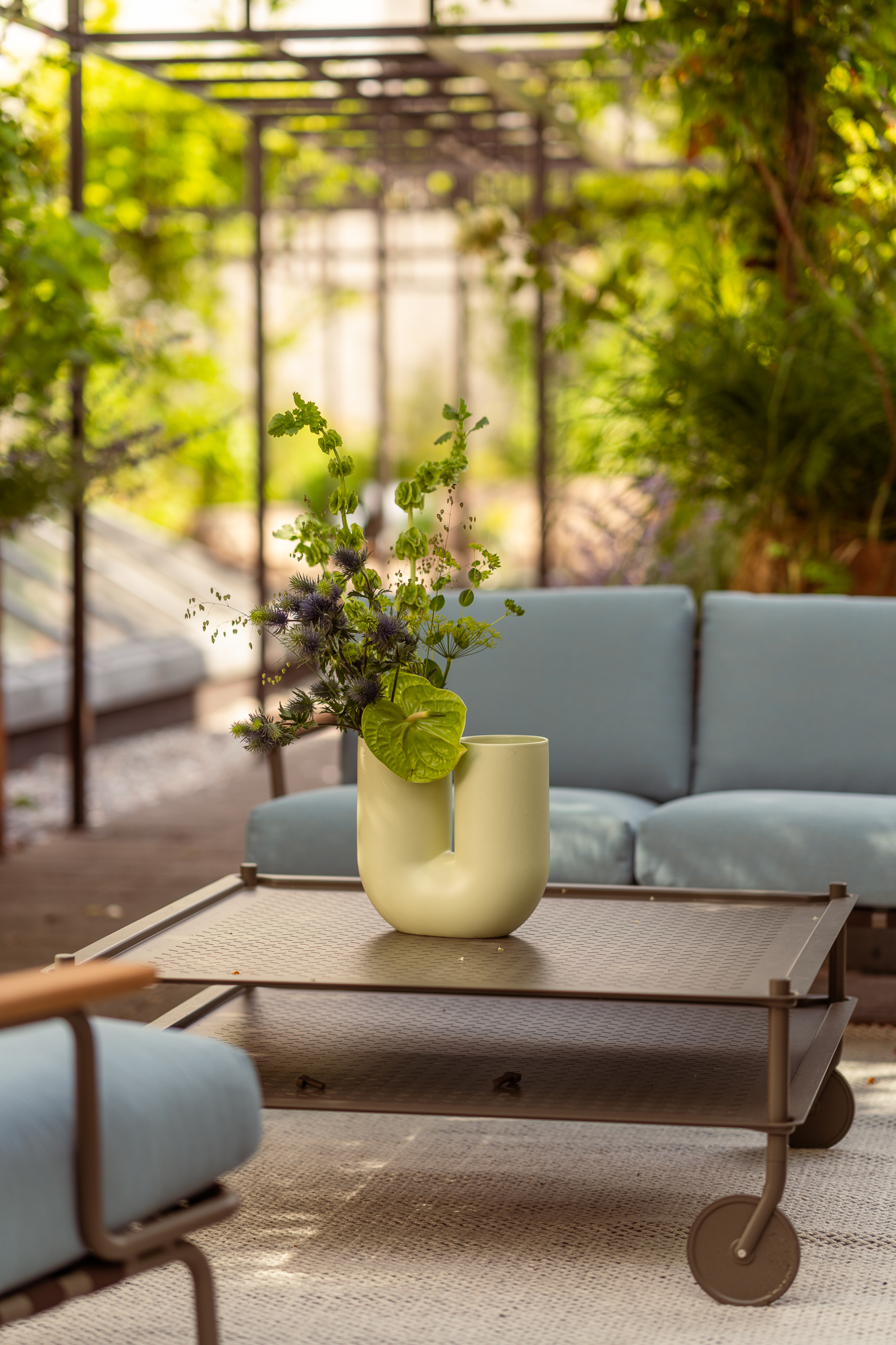 A light blue Settle sofa and matching chair are arranged in a sunny courtyard, surrounded by lush green plants. The seating ensemble rests on a light beige rug, complemented by an outdoor table, creating a cozy and inviting atmosphere.