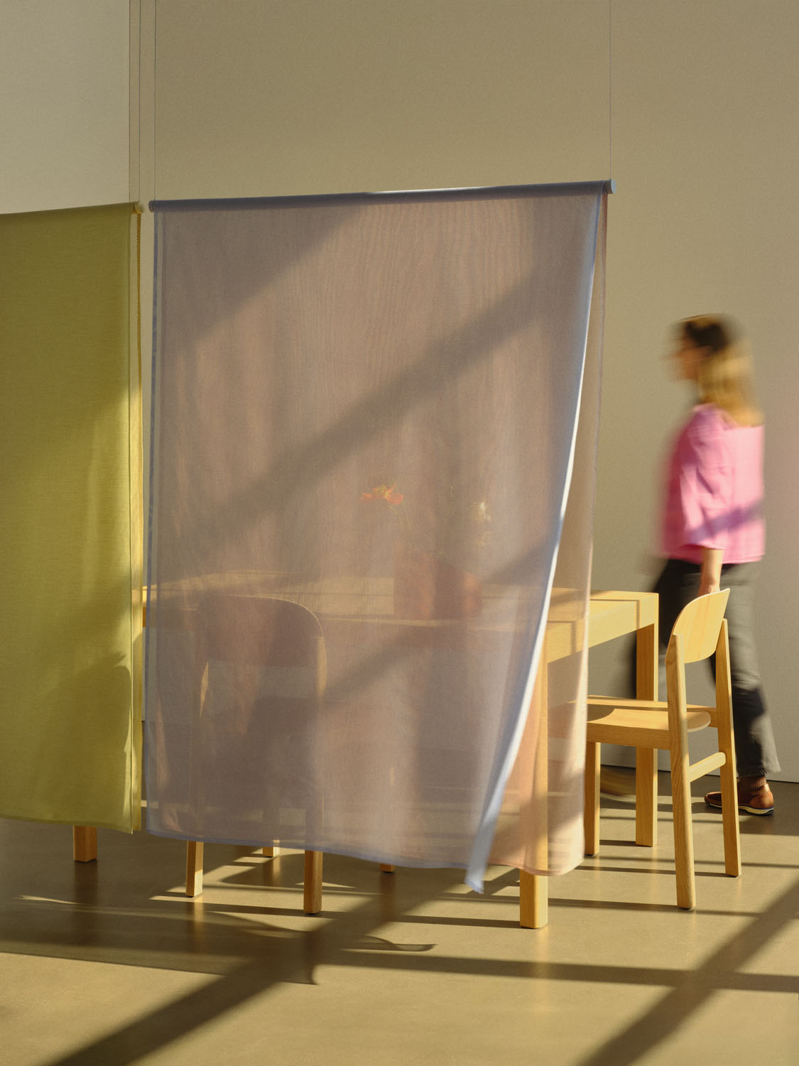 Full view of a softly lit space where two “Veil” panels—green and lavender—hang from the ceiling to divide a minimalist wooden dining setup. A person in pink walks by the panels, adding scale and reinforcing the project’s aim to softly delineate interior zones without enclosing them.

