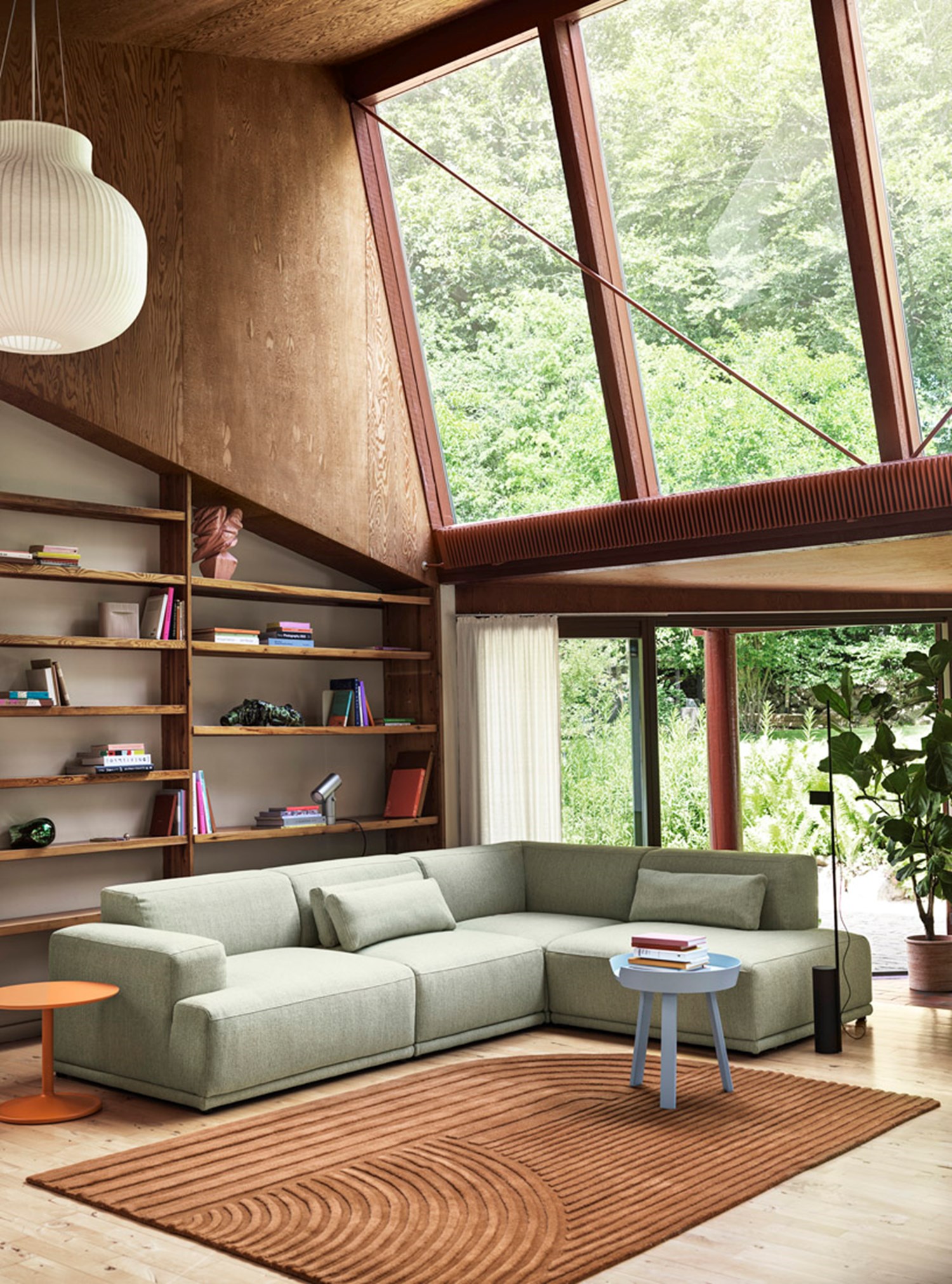 A modern living room with large windows showcasing a lush green forest view. The room includes a light green Connect Soft Modular Sofa, a small blue side table, and a white ribbed pendant light, creating a serene reading corner. A modern living room with large windows showcasing a lush green forest view. The room includes a light green Connect Soft Modular Sofa, a small blue side table, and a white ribbed pendant light, creating a serene reading corner.