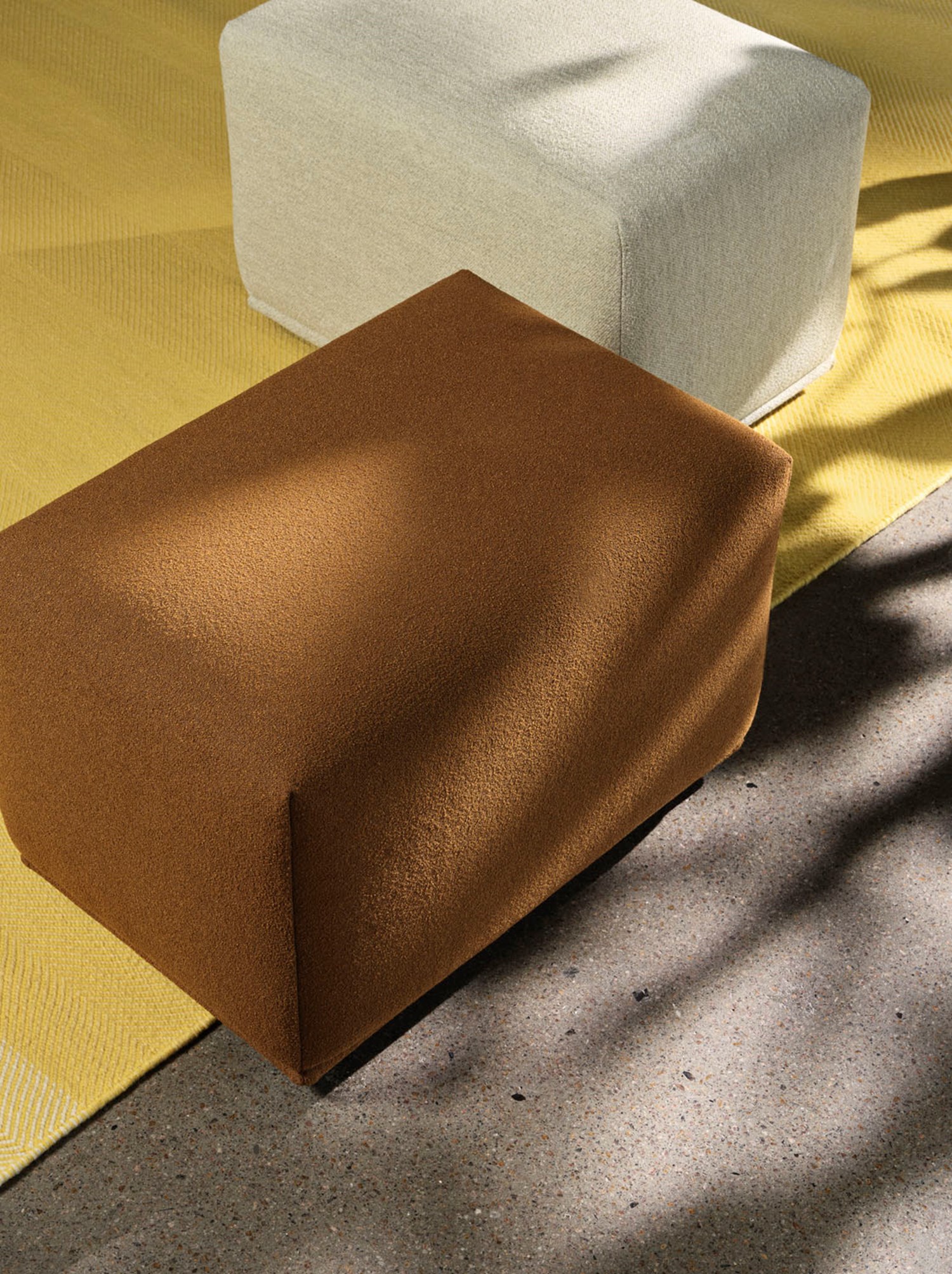 A geometric tan-colored ottoman and a light gray fabric block on a textured yellow rug, highlighted by natural sunlight. A geometric tan-colored ottoman and a light gray fabric block on a textured yellow rug, highlighted by natural sunlight.